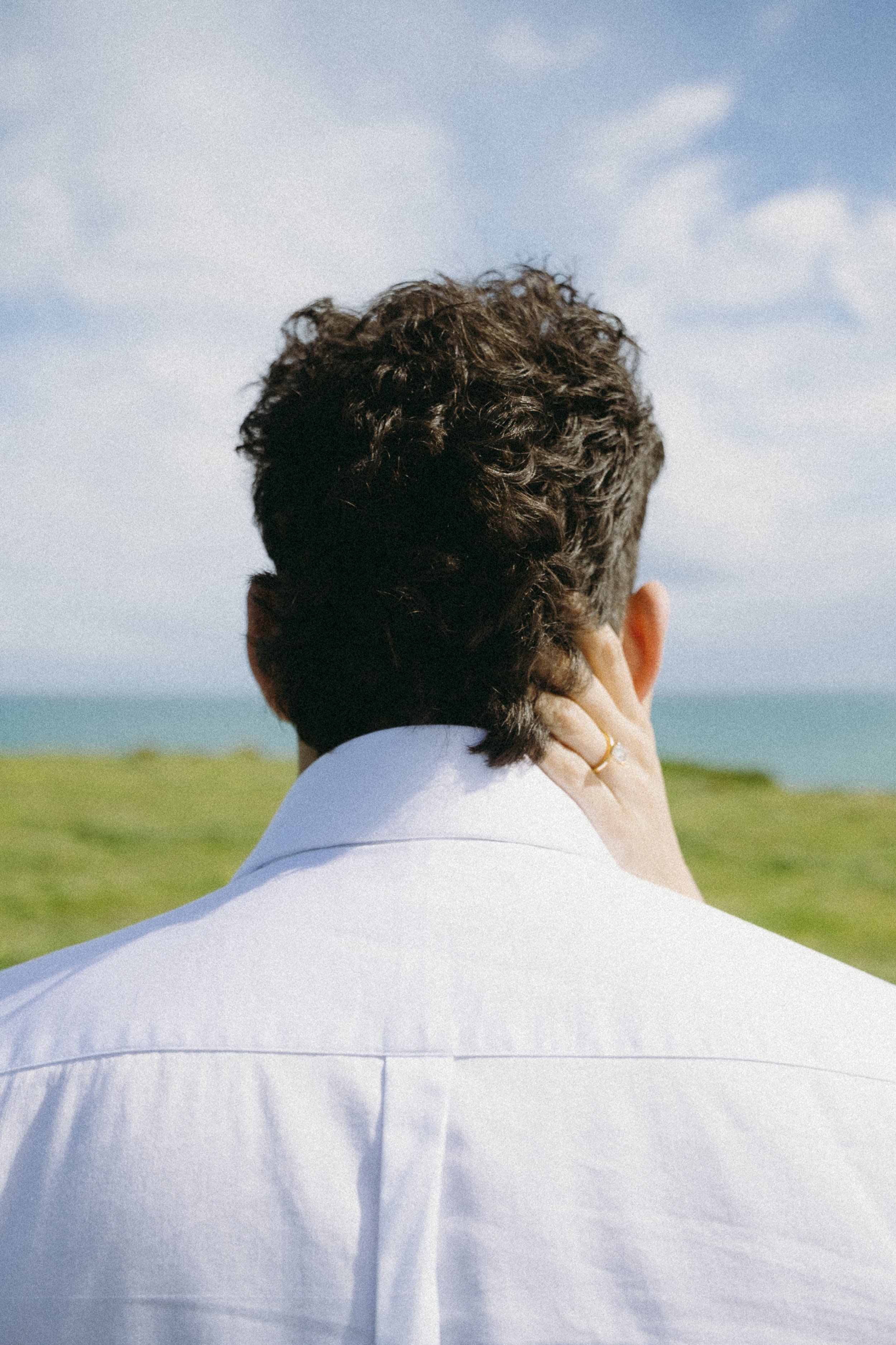 Back view of a man with curly hair wearing a white shirt, standing outdoors with grassy landscape and blue sky in the background.