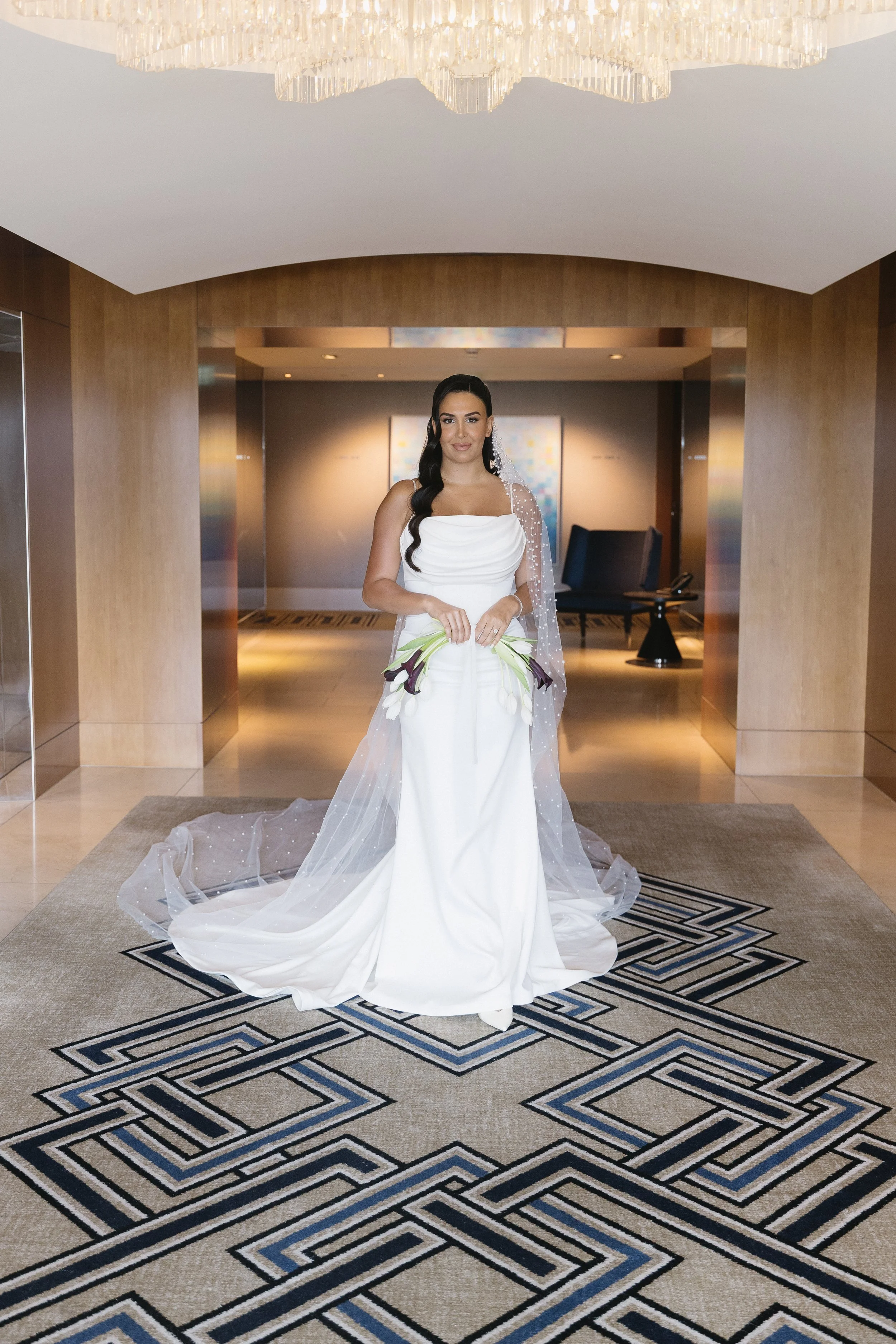 A bride in a white wedding dress holding calla lilies stands in a hotel lobby with wood paneling, modern artwork, and patterned carpet.