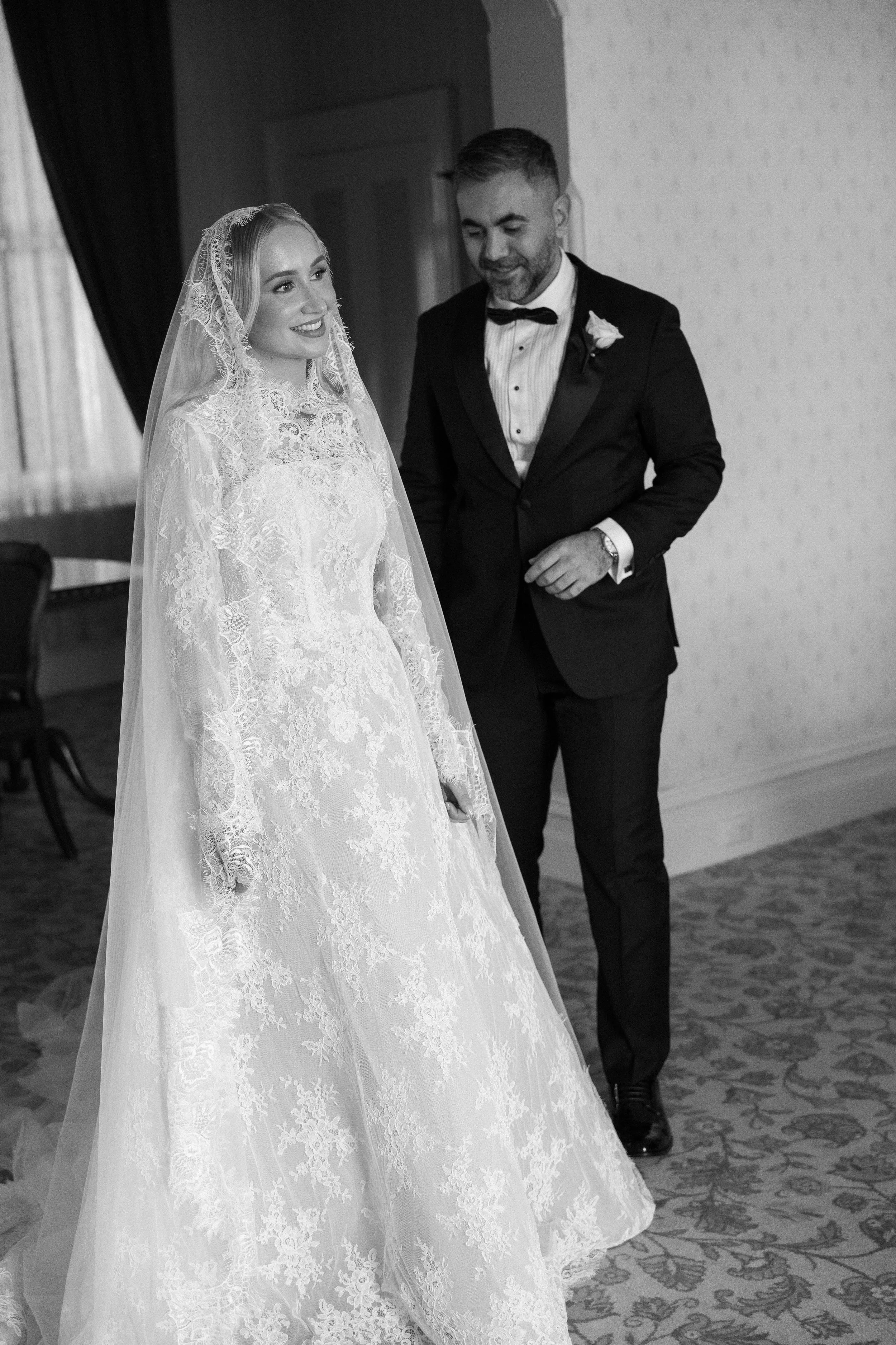 A bride in a lace wedding dress and veil standing next to a groom in a tuxedo indoors.