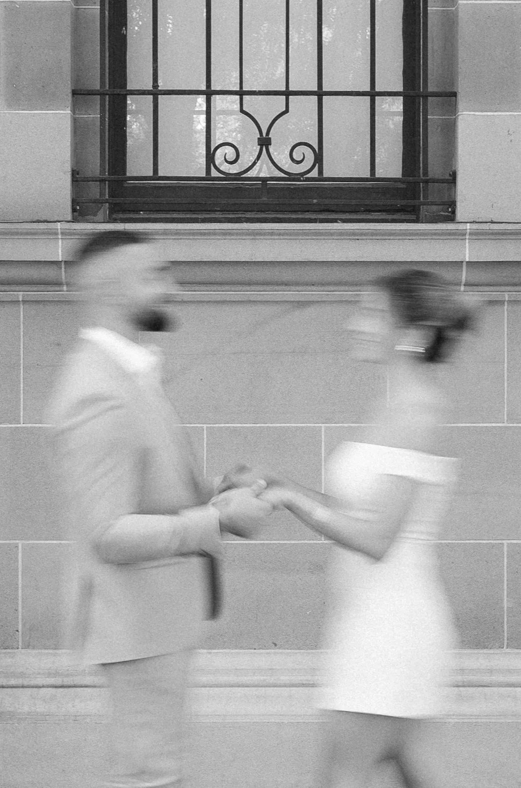 A black-and-white photo of a man and woman holding hands and facing each other, with a building and a decorative window in the background. The image is blurred, emphasizing movement.