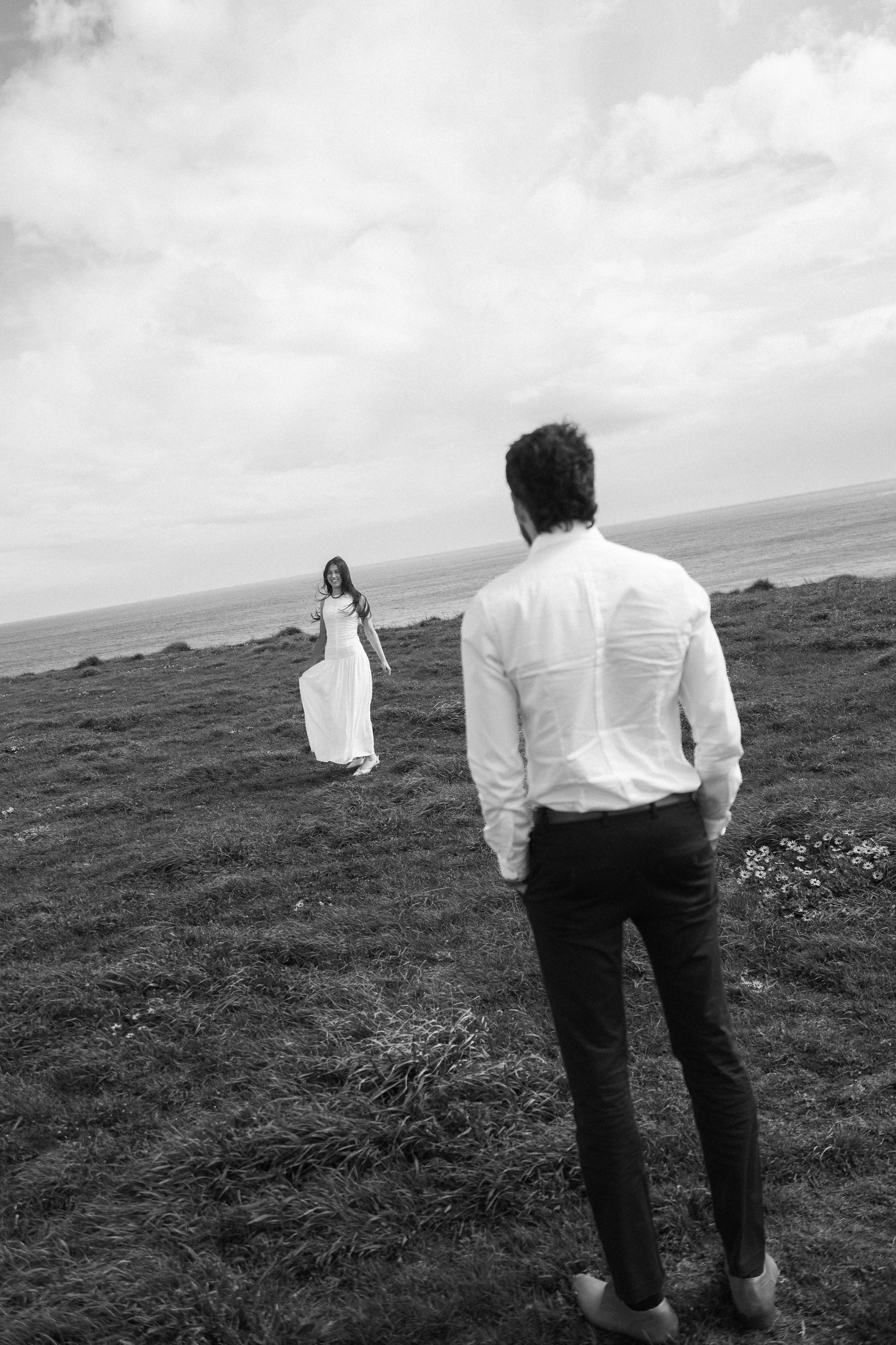 A man in a white shirt and dark pants stands on a grassy field, looking at a woman in a long white dress, who is walking towards him with the sea and cloudy sky in the background.