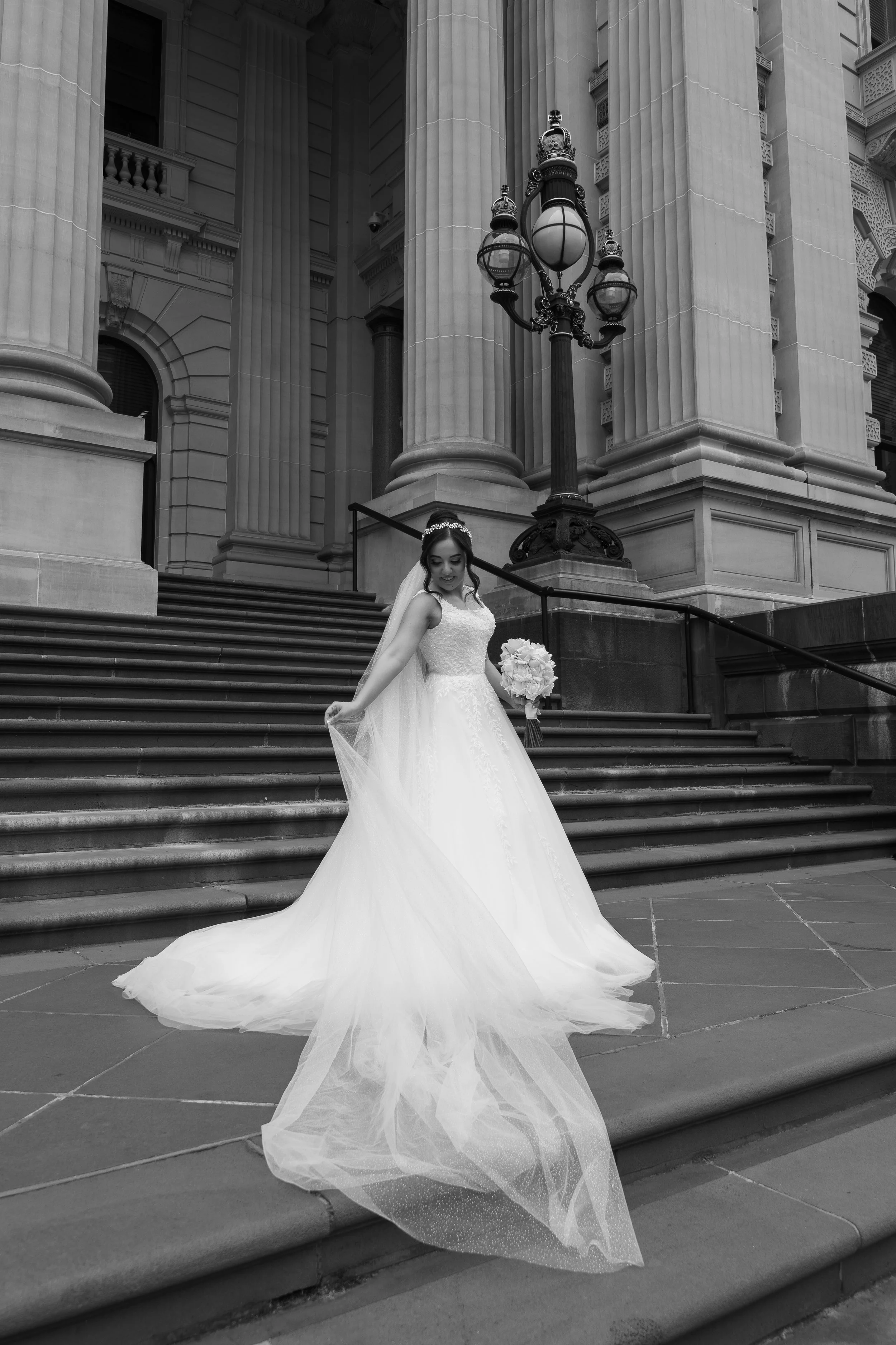 A bride in a wedding dress holding a bouquet, standing on stairs in front of a building with large columns and a decorative streetlamp.