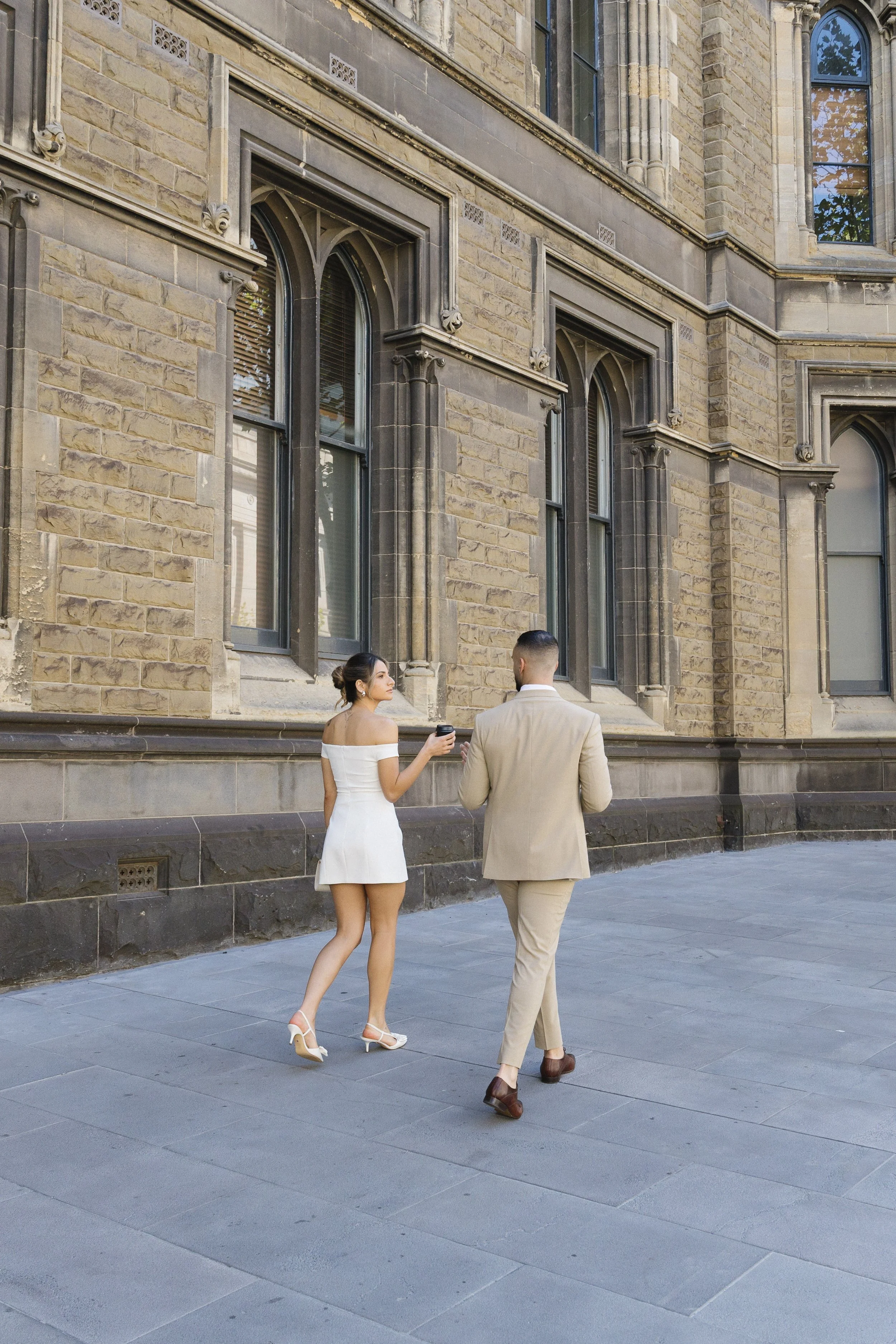 A woman in a white off-shoulder dress and high heels is walking with a man in a beige suit and brown shoes on a city sidewalk in front of a historic brick building with large arched windows.