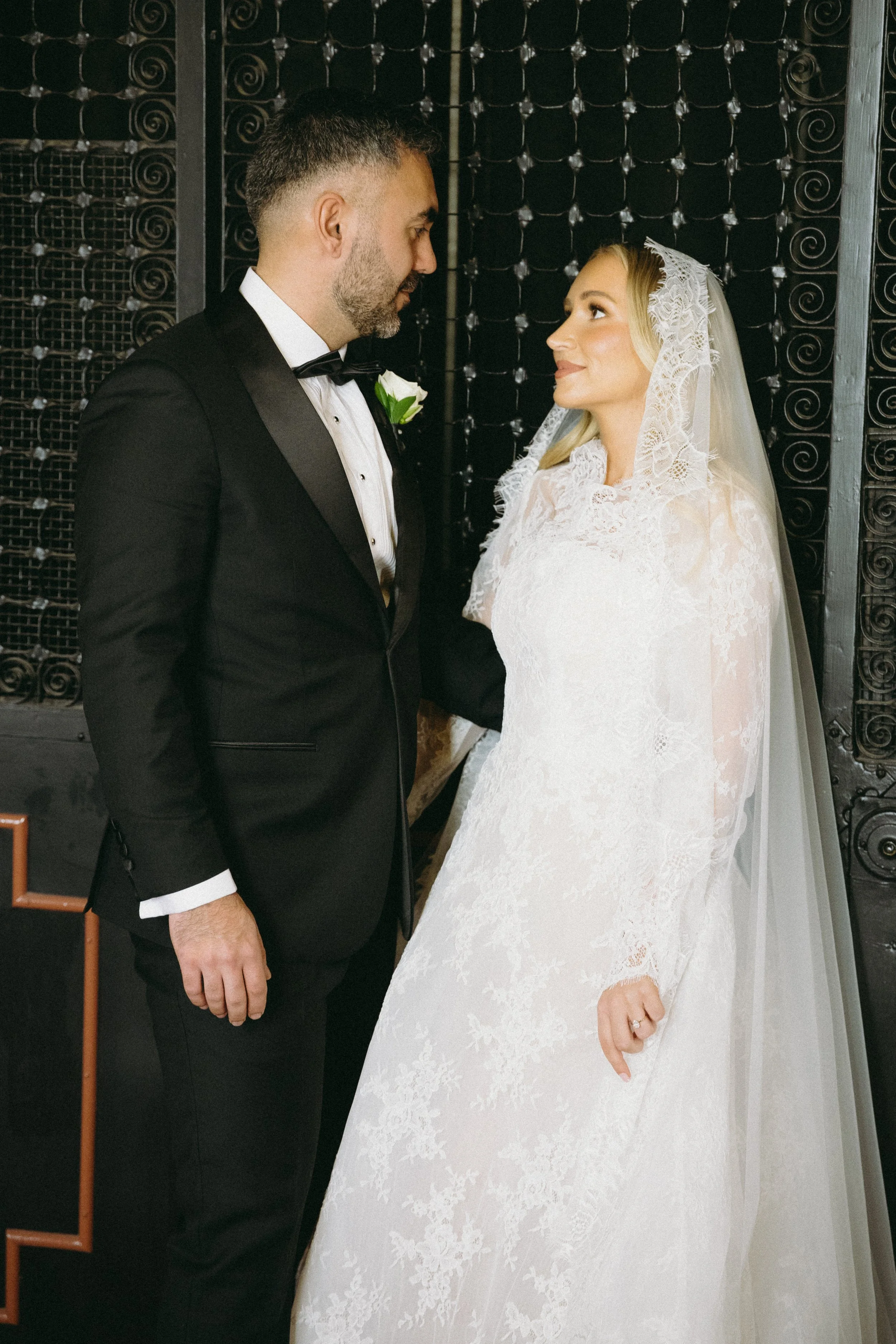 A groom in a black tuxedo with a white shirt and black bow tie stands facing a bride in a white lace wedding gown with a lace veil, holding hands and gazing at each other.