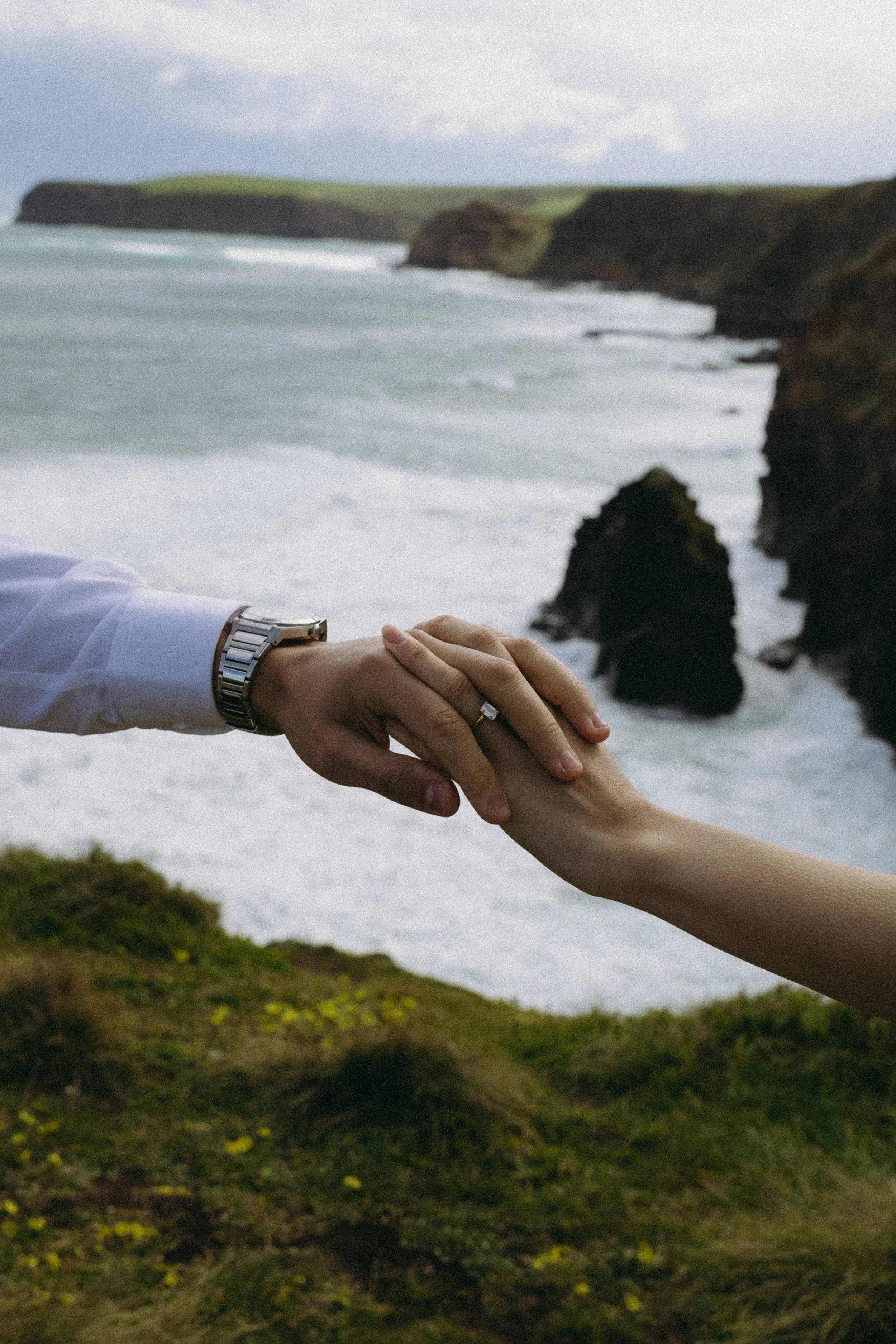 A close-up of a couple holding hands outdoors near the ocean with cliffs and waves in the background.