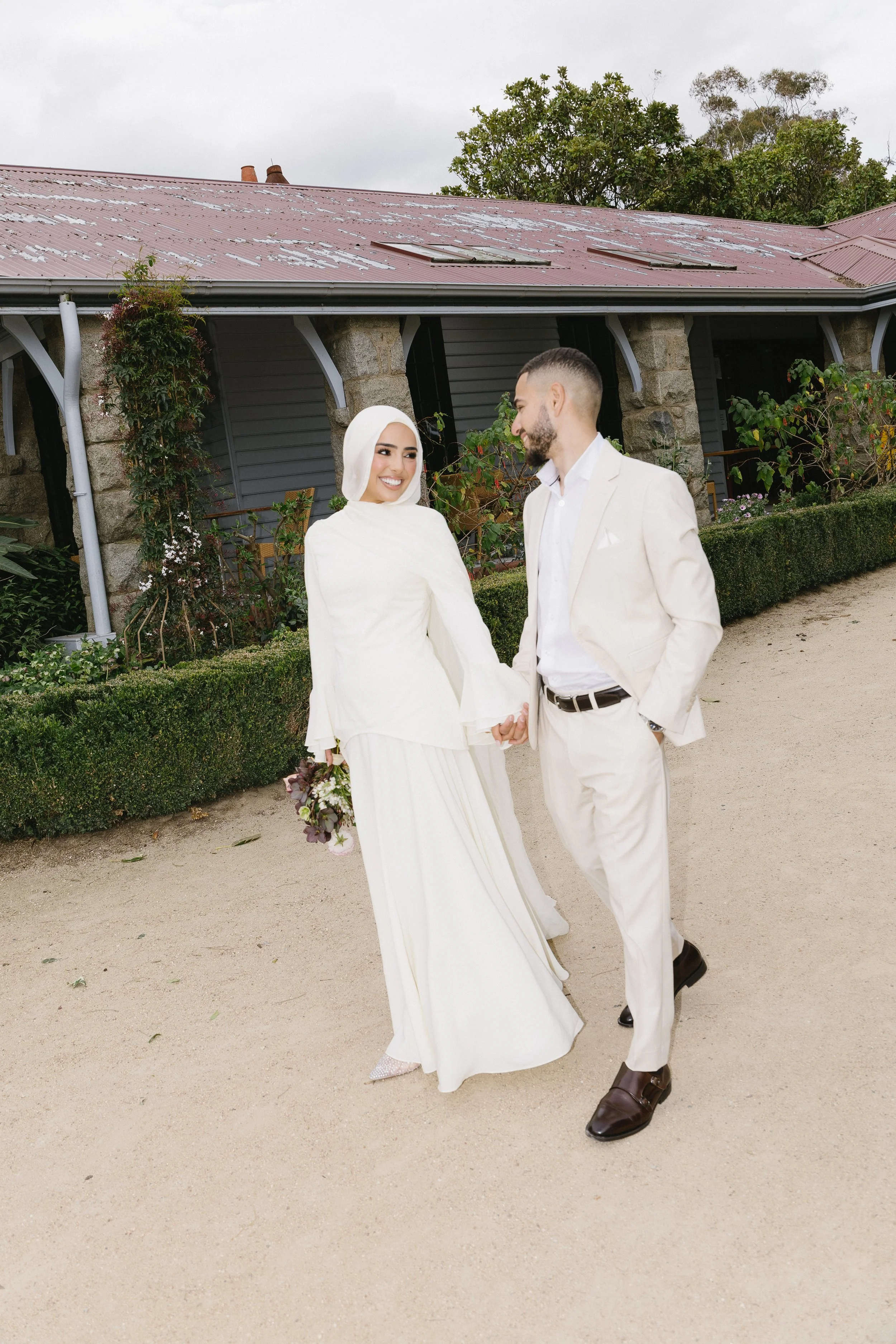 A bride and groom holding hands outdoors, dressed in white wedding attire, standing on a dirt path with greenery and a building with a red roof in the background.