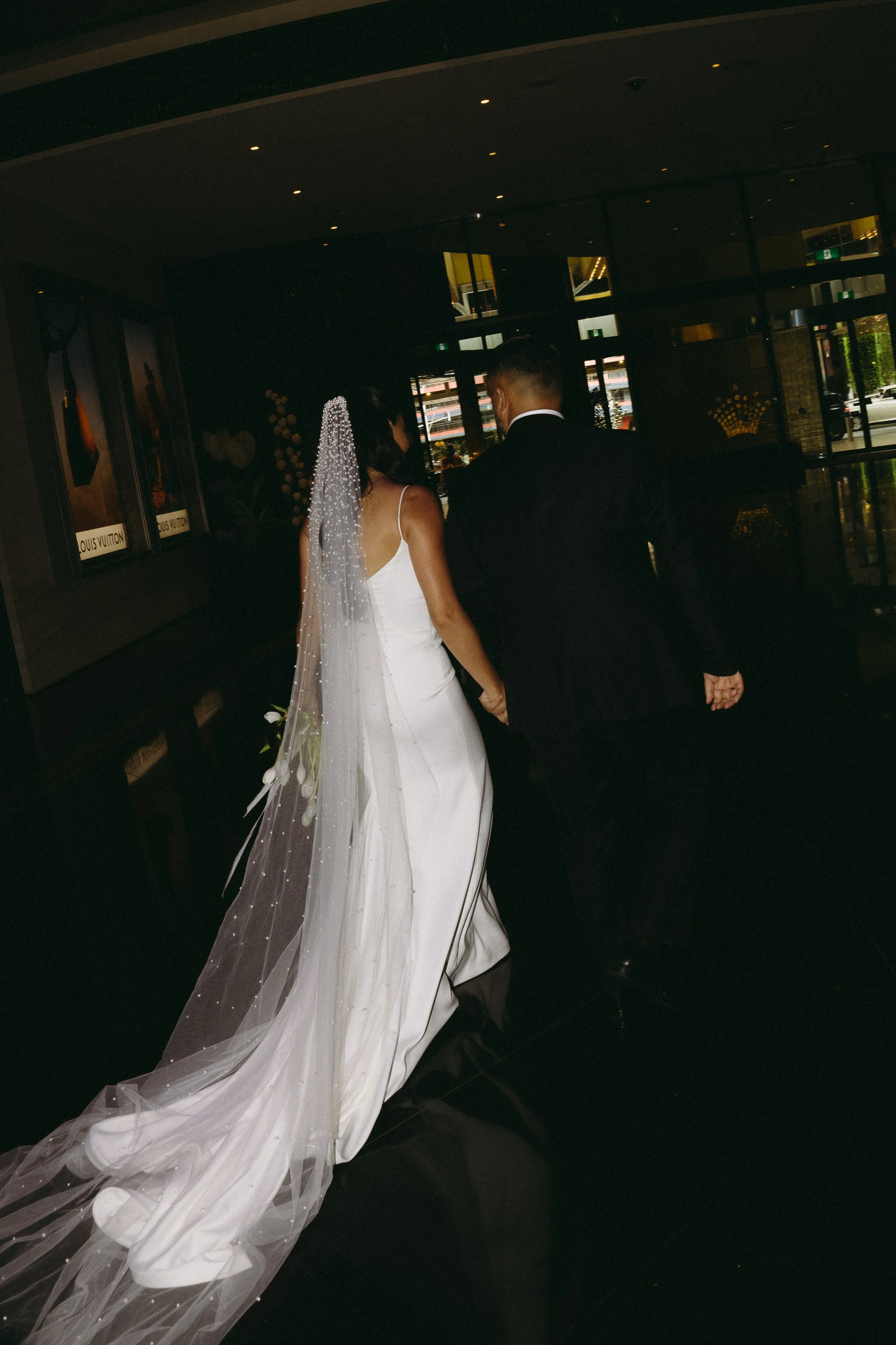 A bride and groom walking hand in hand in a dimly lit indoor space, with the bride wearing a white wedding gown and veil, and the groom in a black suit.