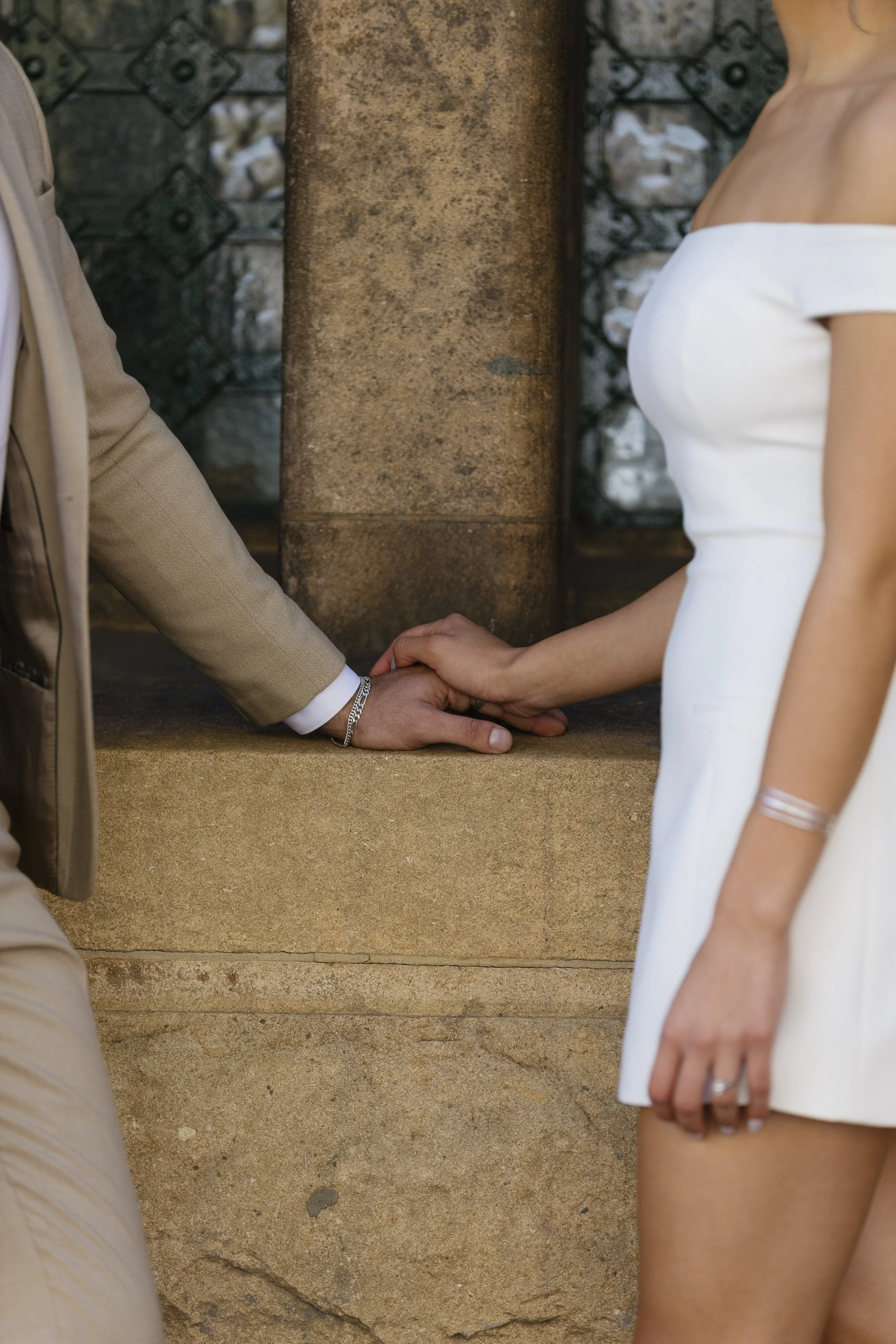 A man and woman holding hands in front of a stone wall, with the woman's hand resting on top of the man's hand.