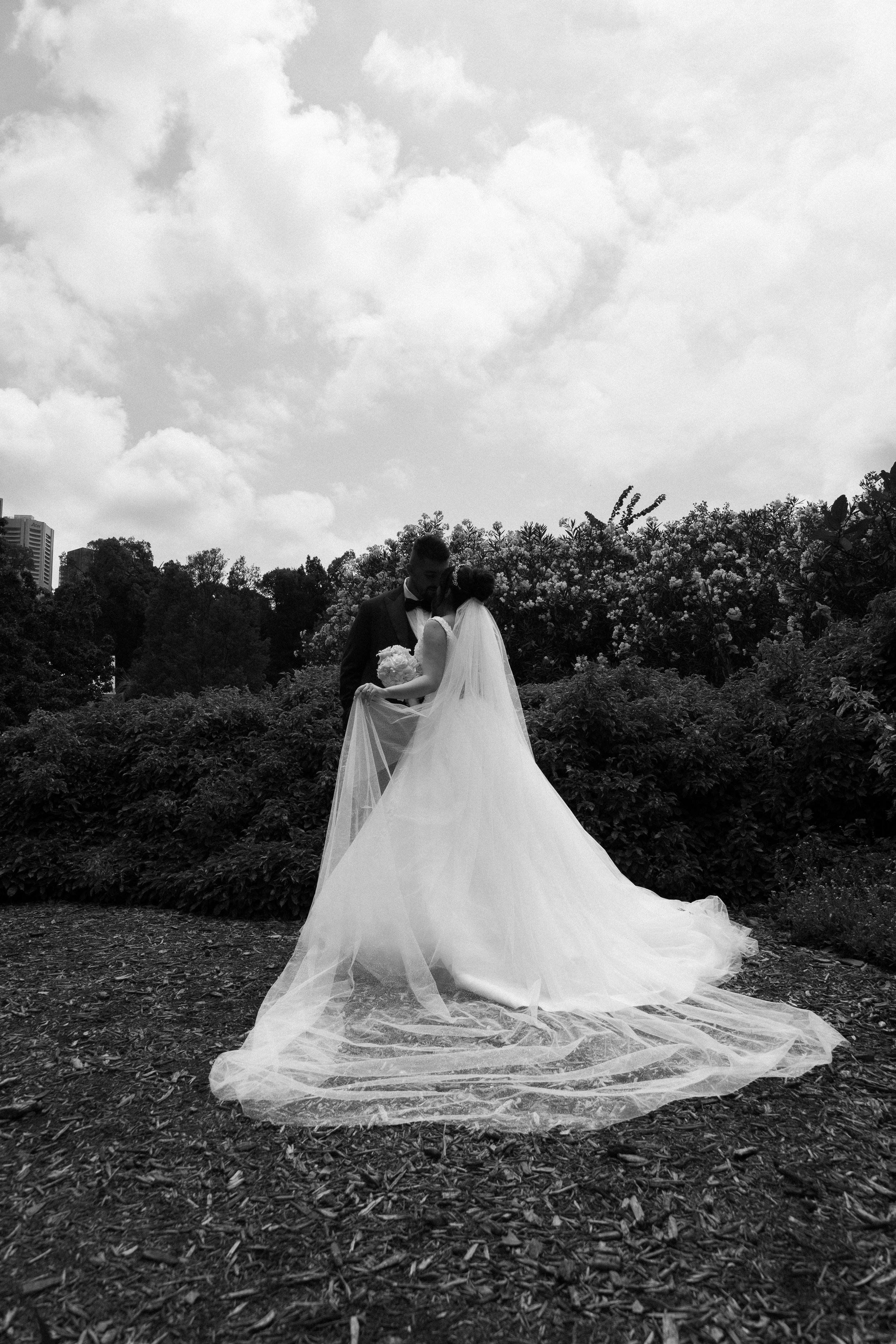 Black and white photo of a bride and groom standing close together outdoors, with the bride wearing a long wedding dress and veil, and the groom in a suit. They are holding a bouquet of flowers, amidst bushes and trees under a cloudy sky.