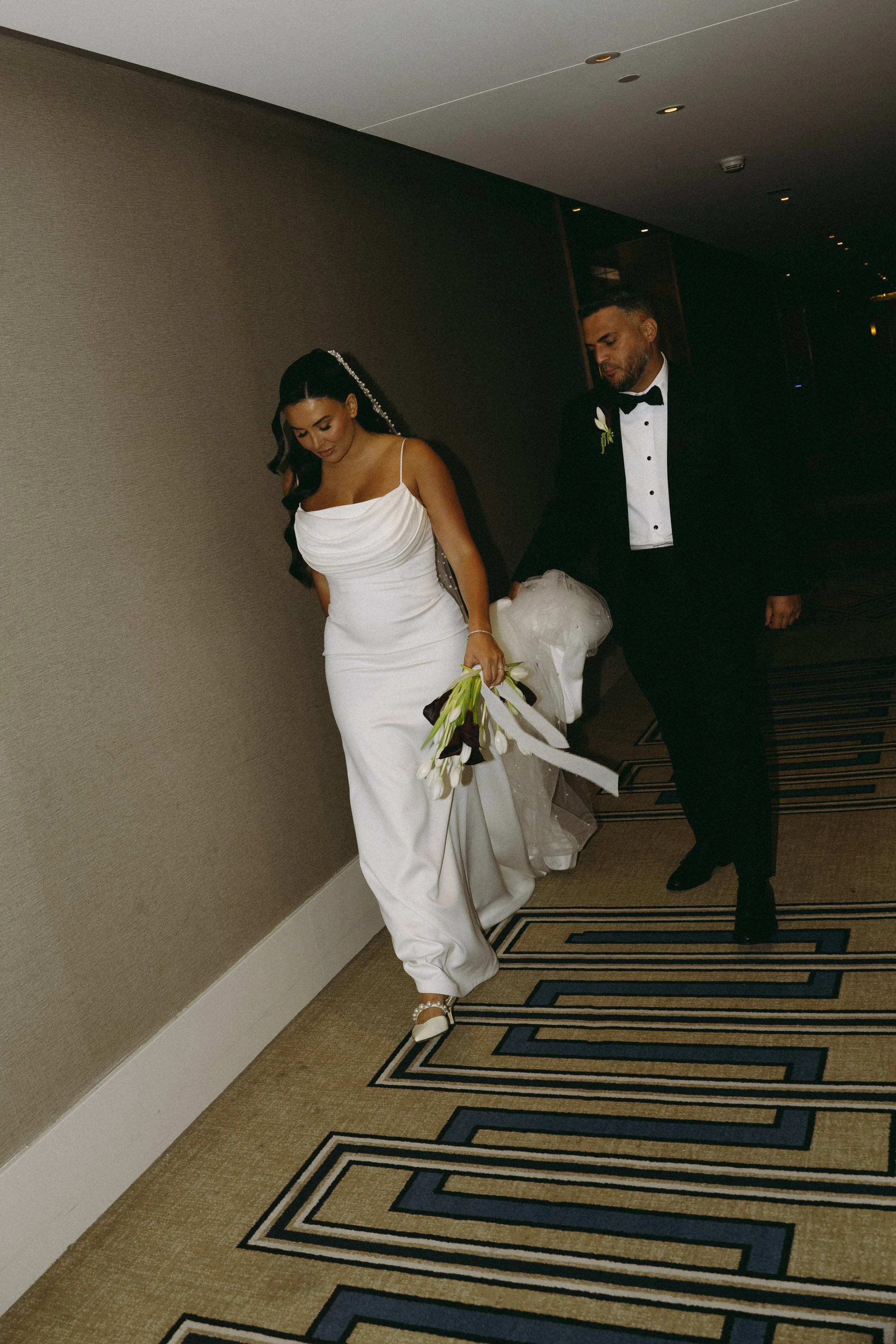 A newly married couple, dressed in formal wedding attire, walking down a hotel corridor. The bride is wearing a white gown, holding a bouquet of white flowers, with a sparkly hair accessory. The groom is in a black tuxedo with a bow tie, holding the train of the bride's dress.