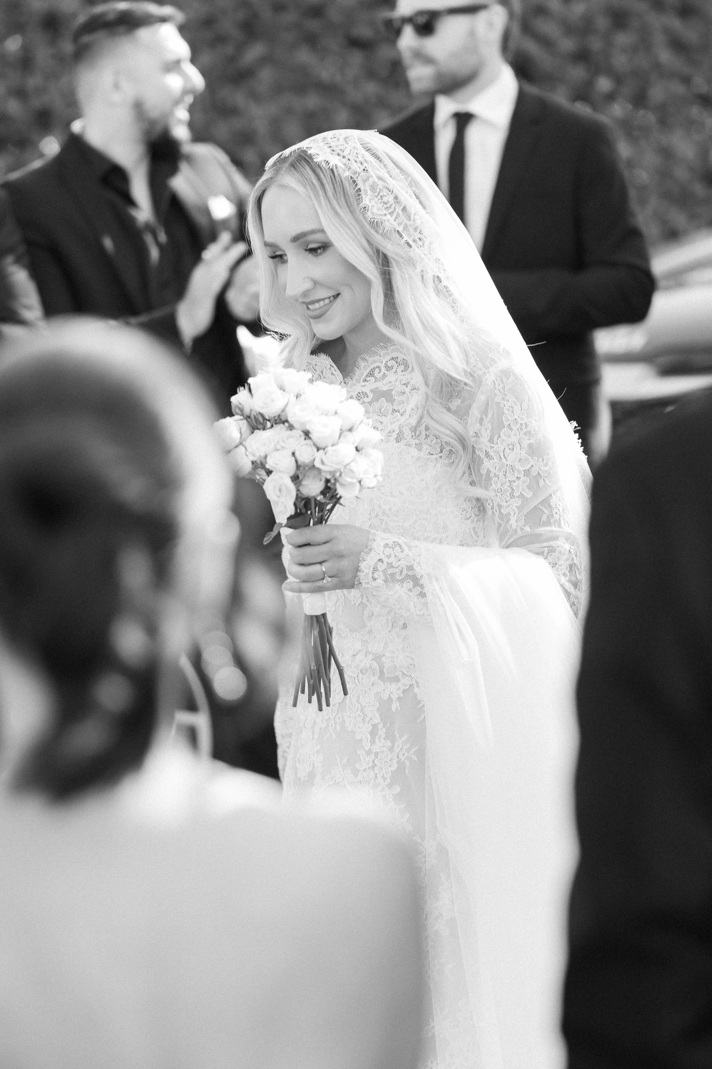 A bride in a lace wedding dress and veil holding a bouquet of flowers, smiling during her wedding ceremony surrounded by guests.