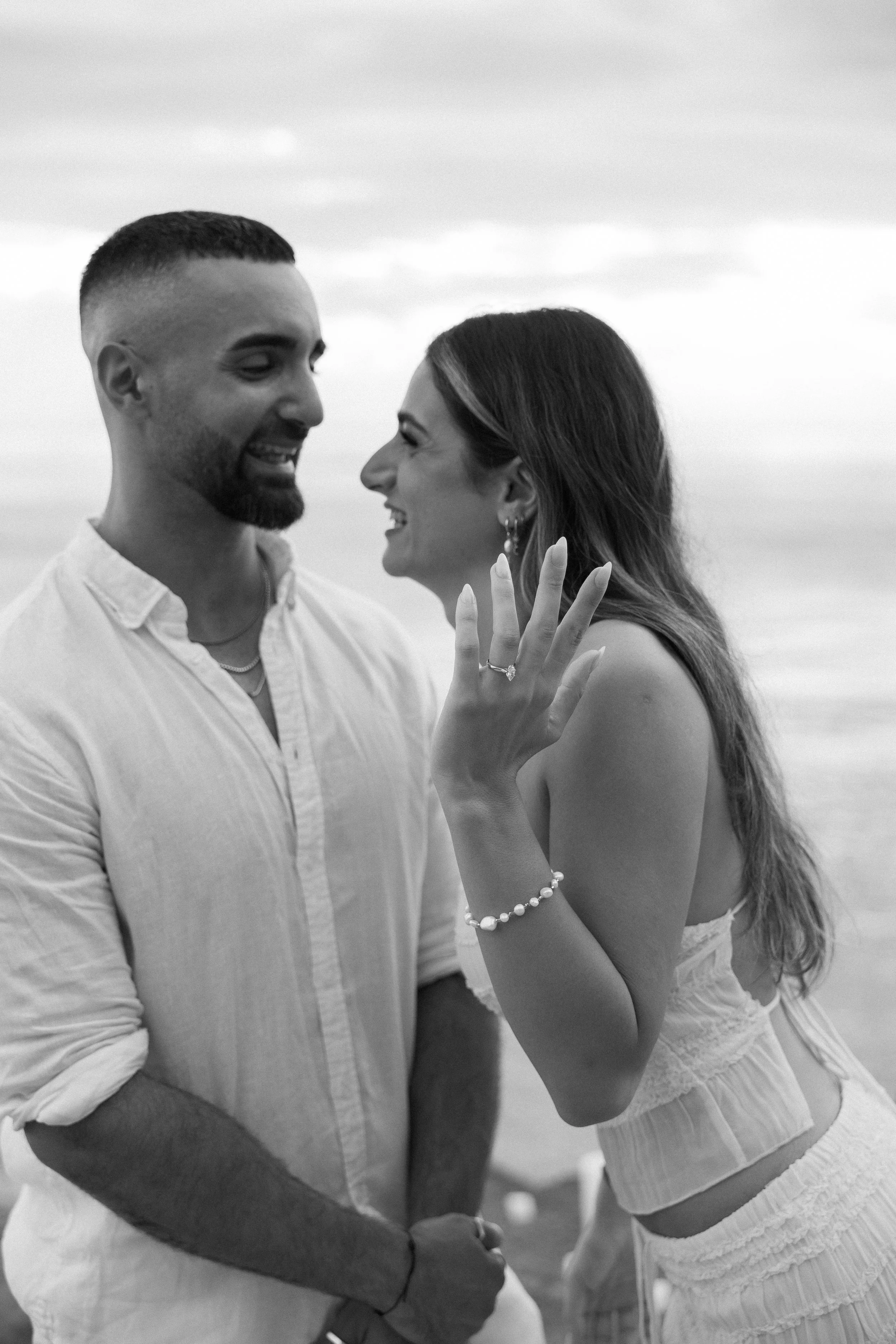A black and white photo of a smiling couple on the beach, with the woman showing off her engagement ring on her left hand.