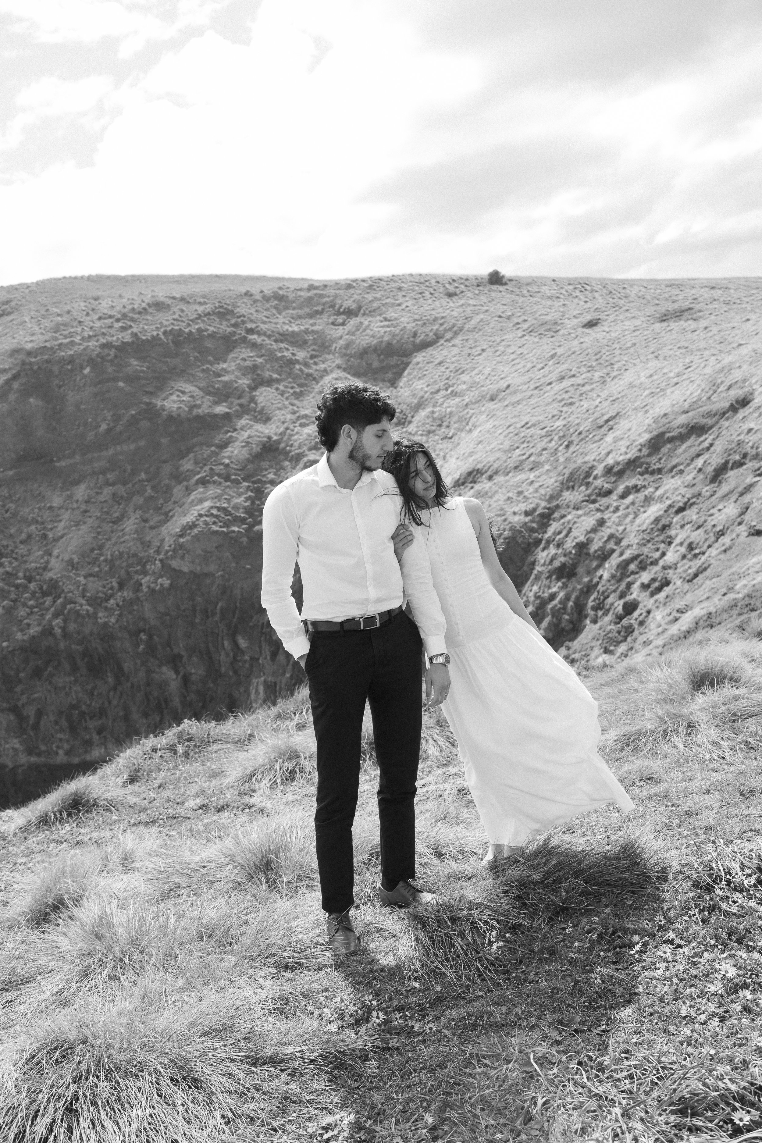 A young couple, a man in a white shirt and black pants, and a woman in a white dress, standing on a grassy hillside with rugged hills in the background. The woman is leaning on the man's shoulder, and they are holding hands, with both looking down or away.
