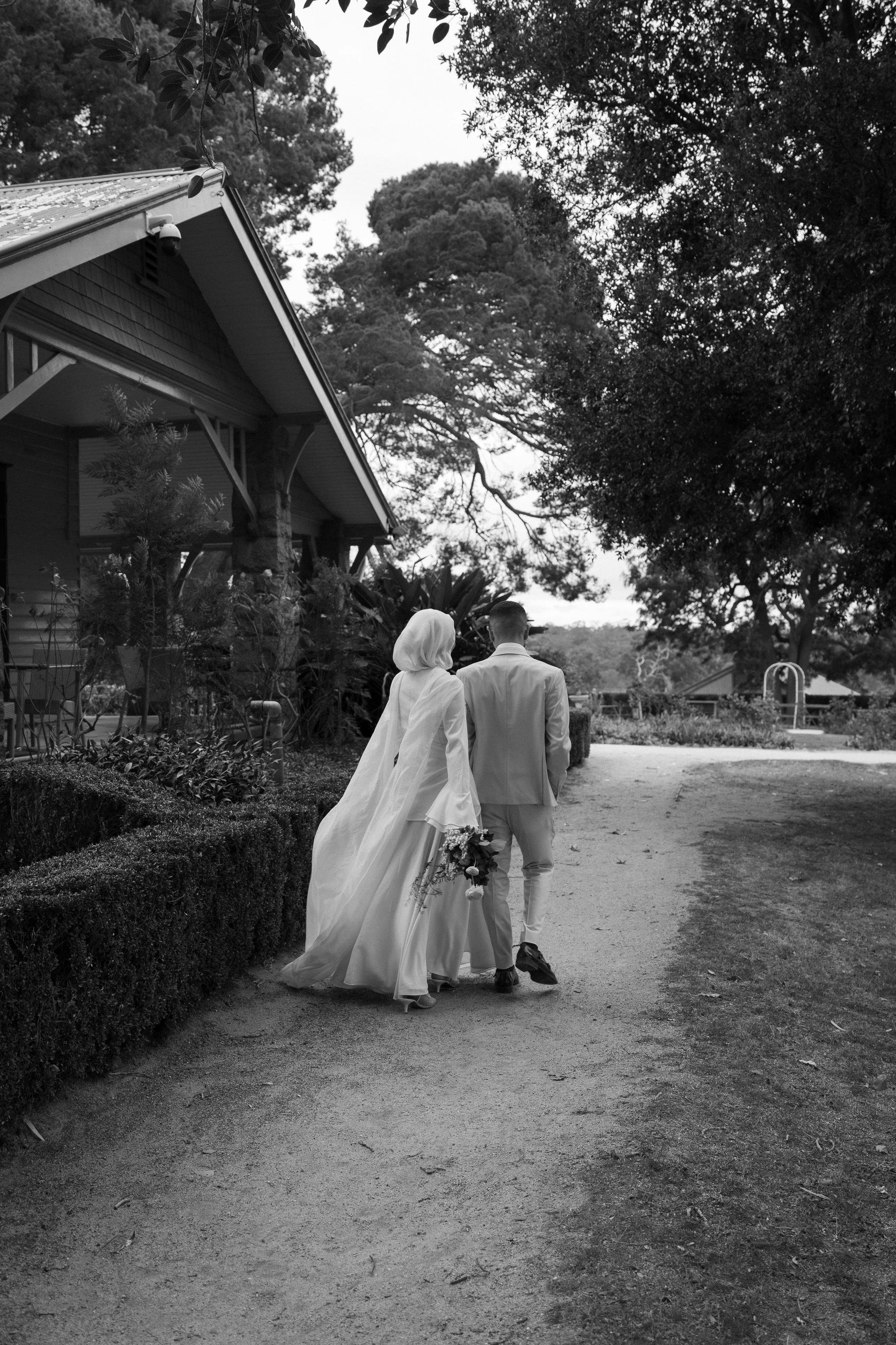 A bride and groom walk together on a path, the bride holding a bouquet, surrounded by trees and a house in the background, in black and white.