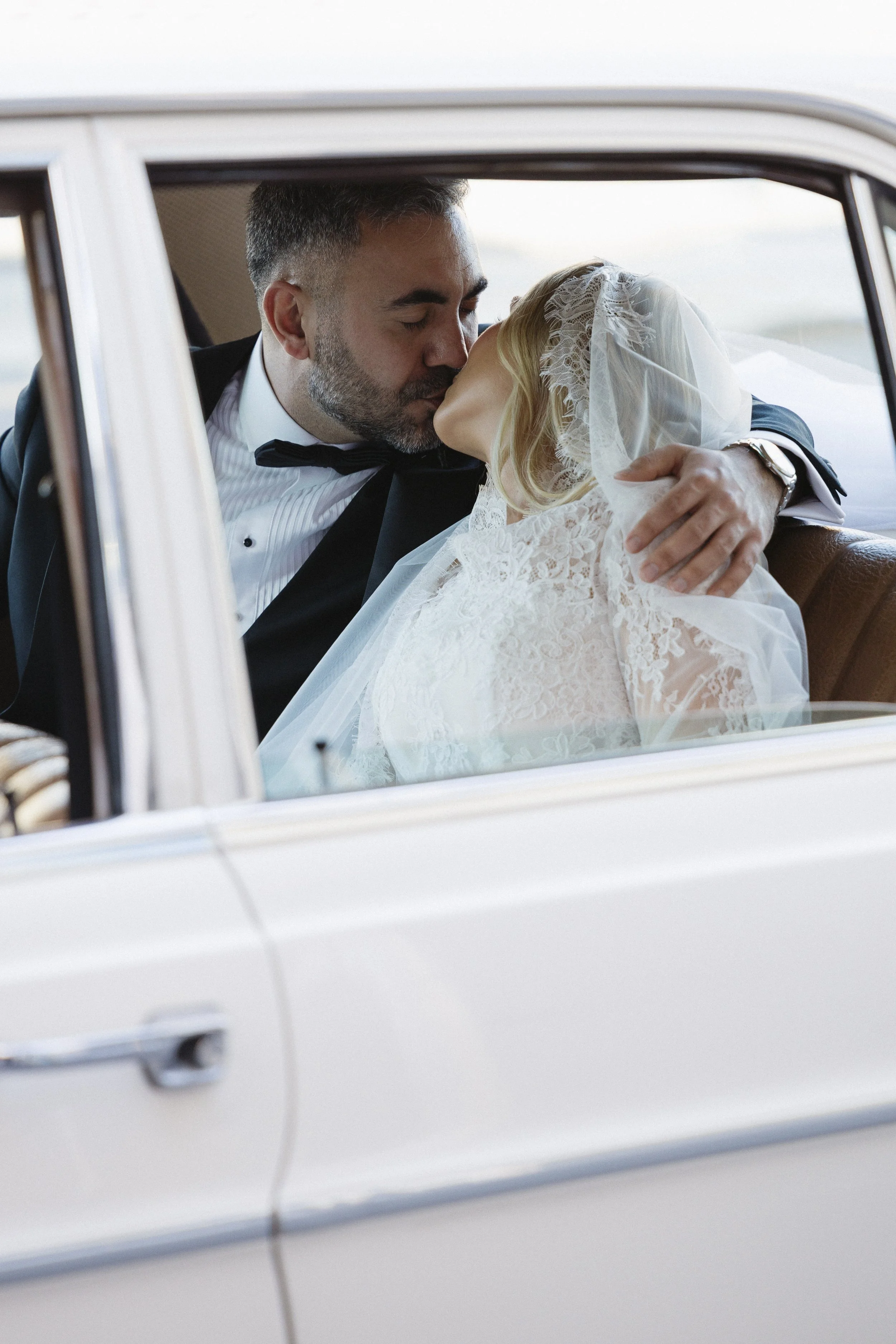 A bride and groom sharing a kiss inside a vintage car, the groom wearing a tuxedo and the bride in a lace wedding dress with a veil.