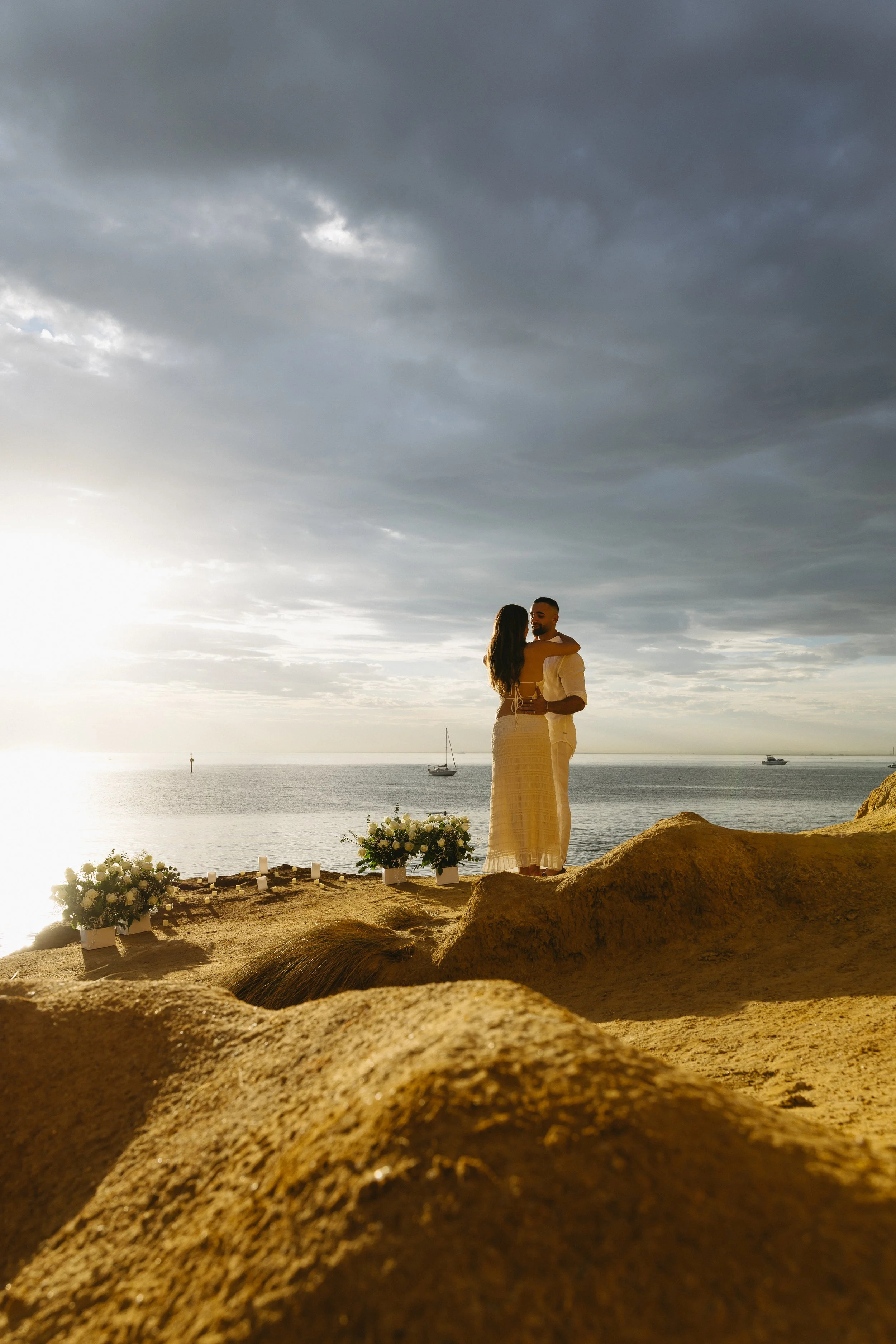A couple embracing on a rocky beach during sunset, with boats on the water and a cloudy sky overhead.
