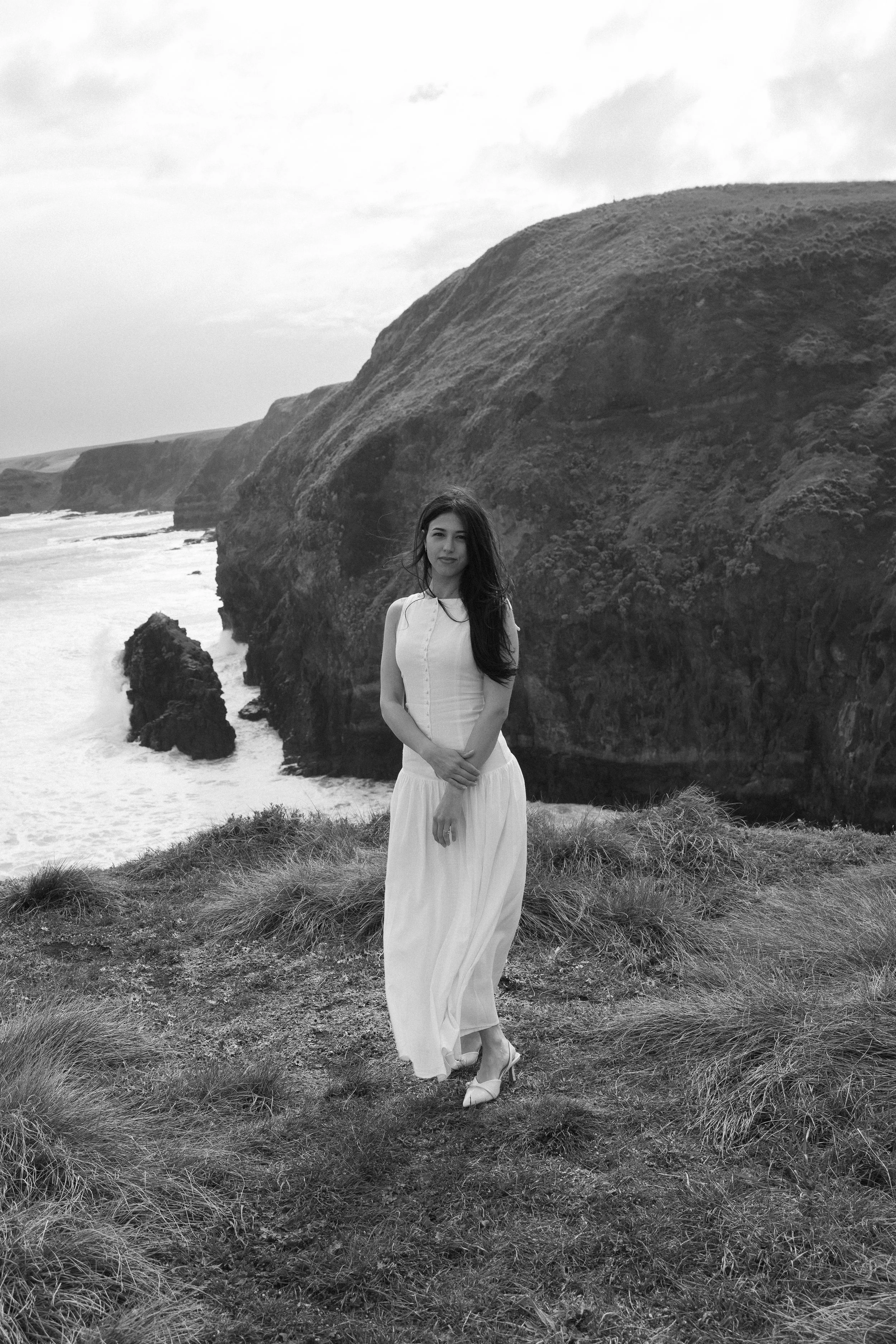A woman in a white dress standing on grass near rocky cliffs with ocean waves crashing below, in black and white.