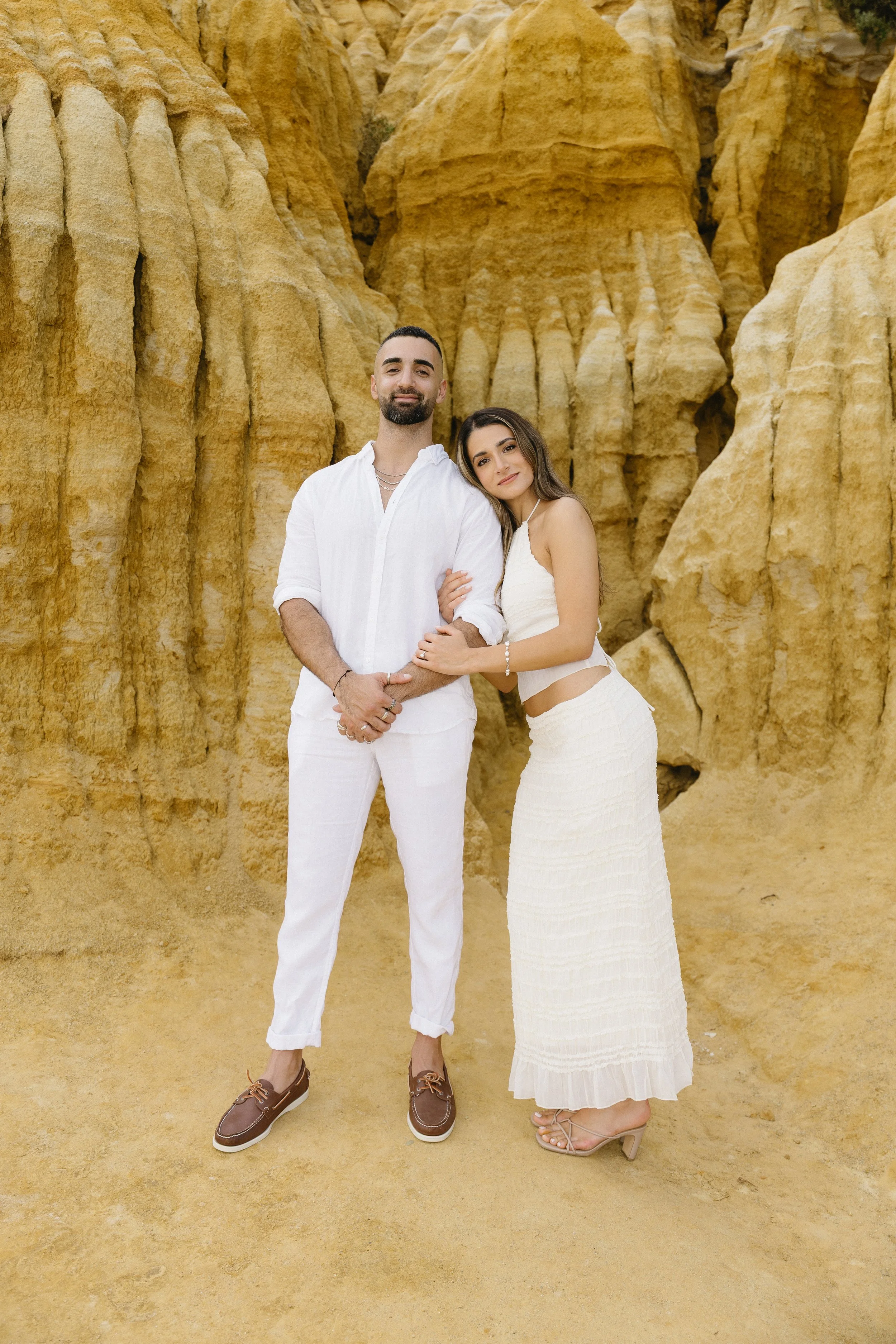 A man and woman stand together in front of yellow rock formations, both dressed in white, with the woman leaning her head on the man's shoulder.