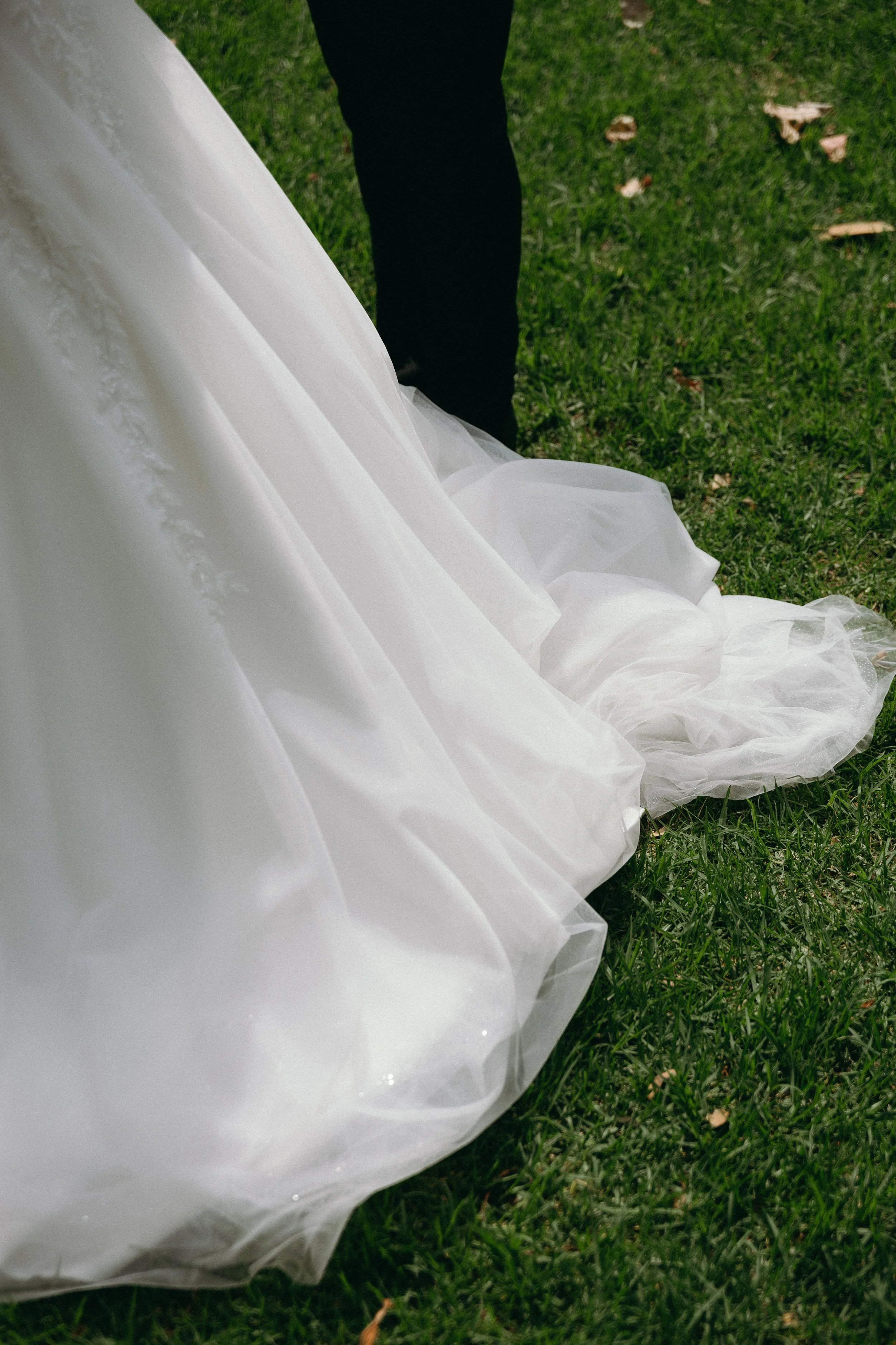 Close-up of a bride's white wedding dress on grass with a groom's black pants visible in the background.