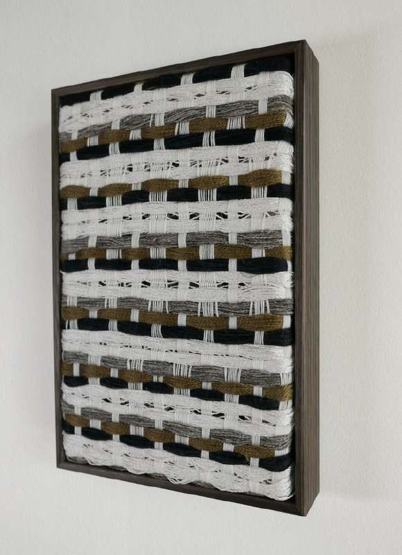 Textile wall art with woven patterns in brown, black, gray, and white, framed in a wooden border hanging on a plain wall.