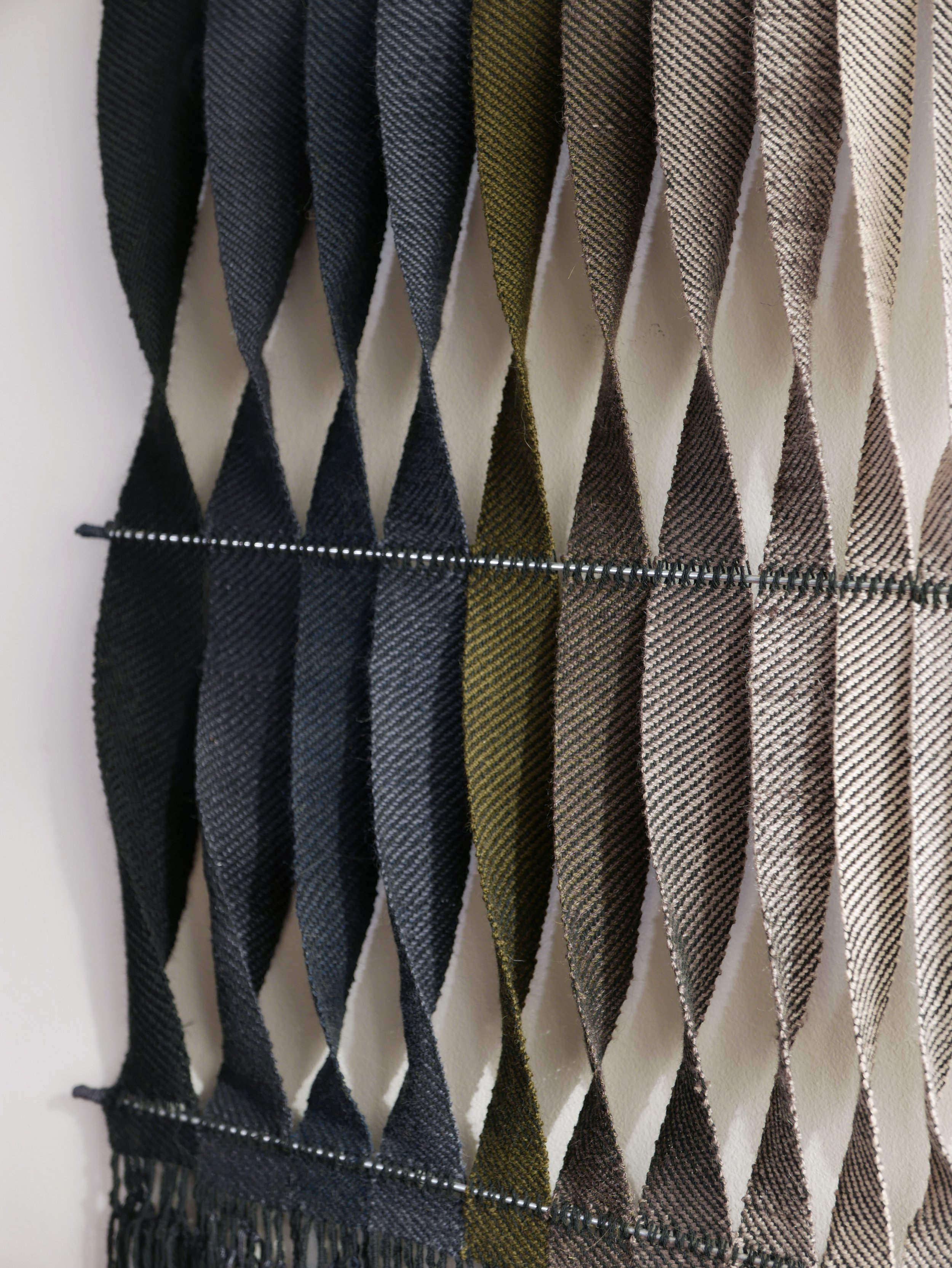 A display of woven neckties hanging on a rack, featuring various colors including black, gray, olive green, dark brown, taupe, and beige, with some ties having a plaid pattern.