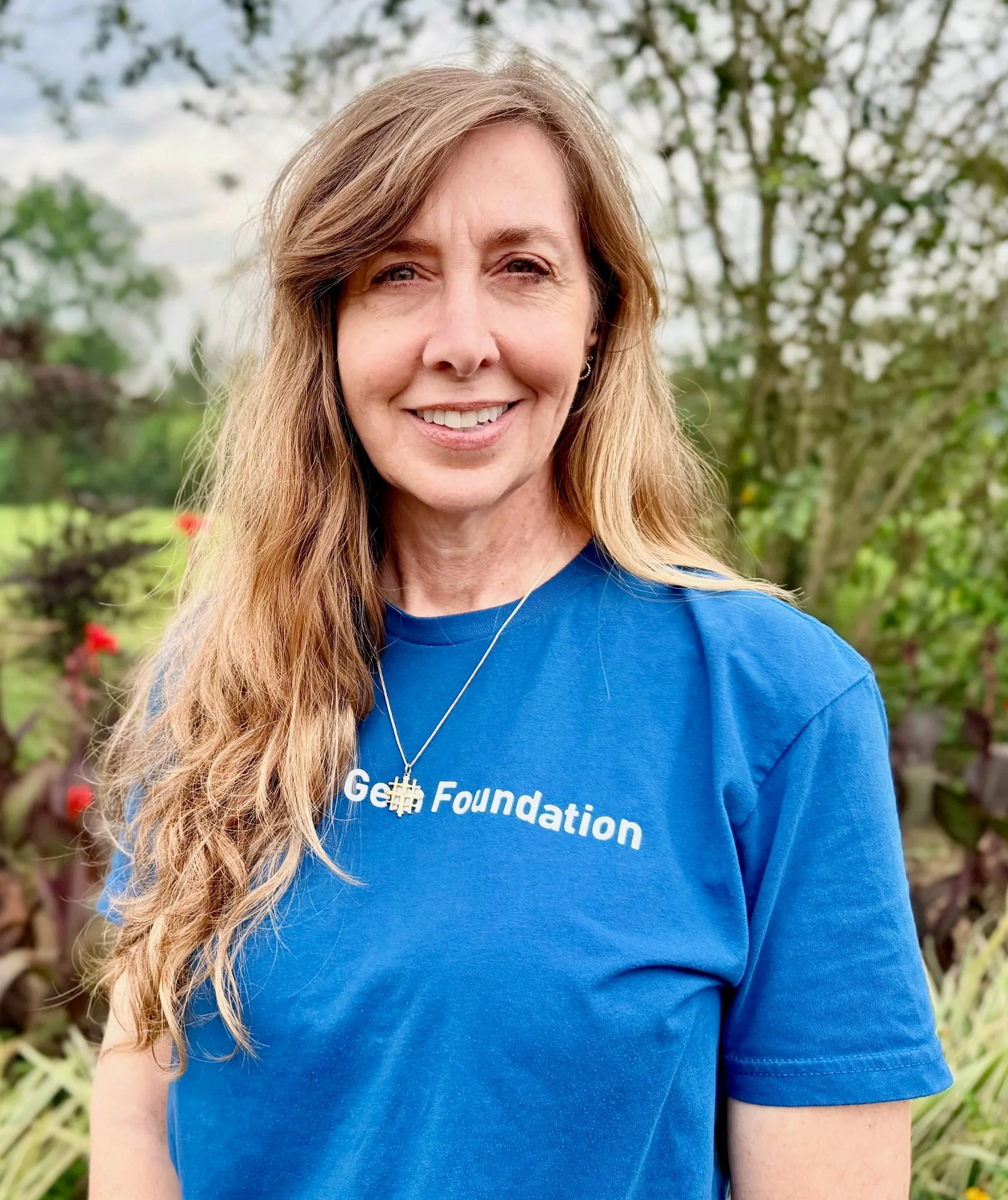 A woman with long blonde hair smiling outdoors in front of green trees, wearing a blue shirt with white text that reads 'The Gem Foundation'.