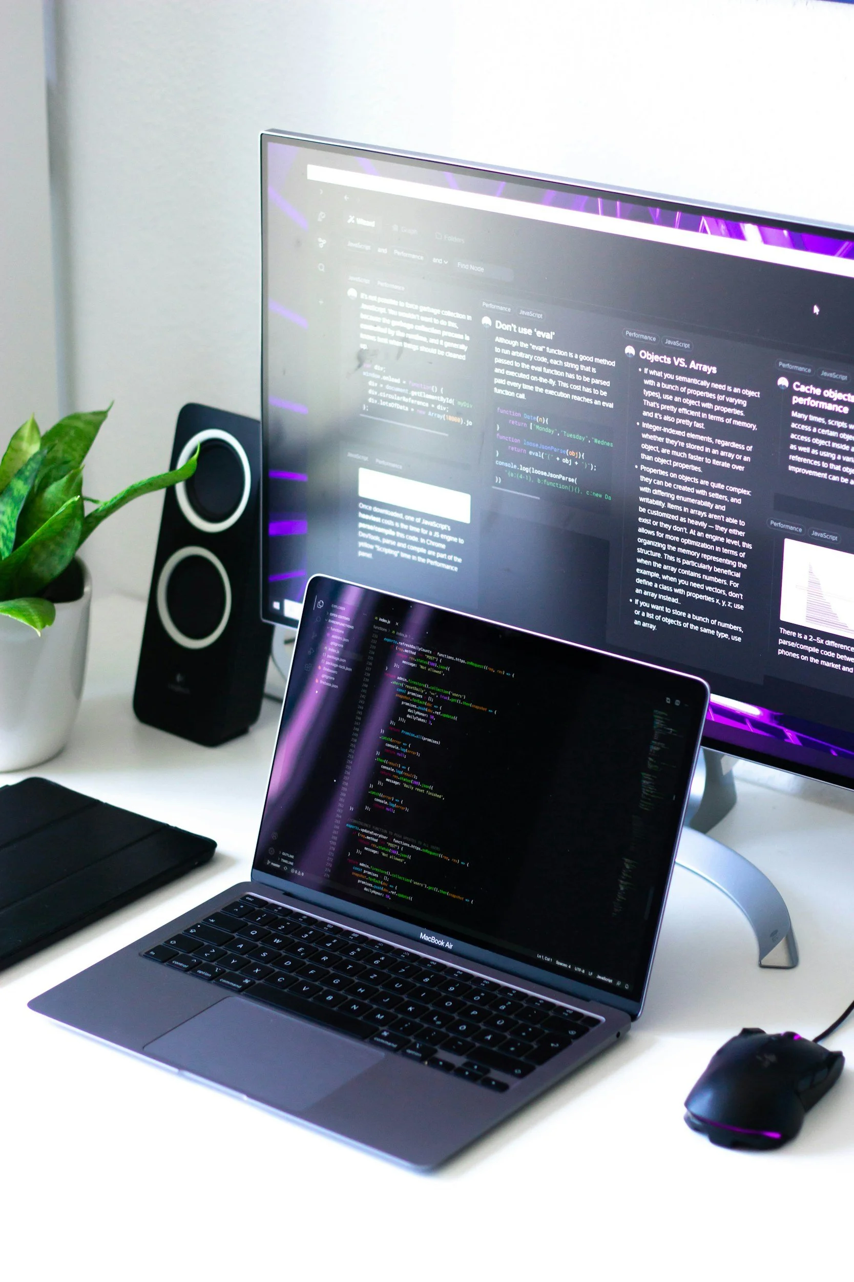 A laptop and computer monitor on a white desk. On the screens is a bespoke software and the coding behind it, the screens have a purple glare about them.