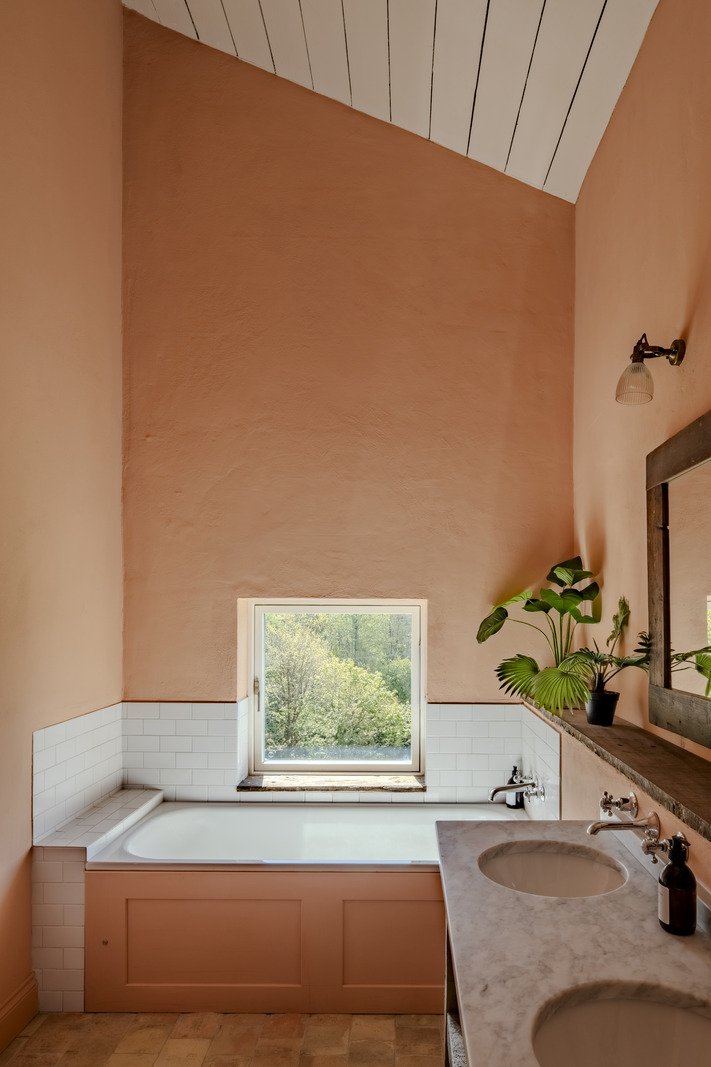 Bathroom with a built-in bathtub beneath a small window, pink walls, white tiled section around the tub, and a marble countertop with two sinks and a large mirror. Green plants sit on the countertop.
