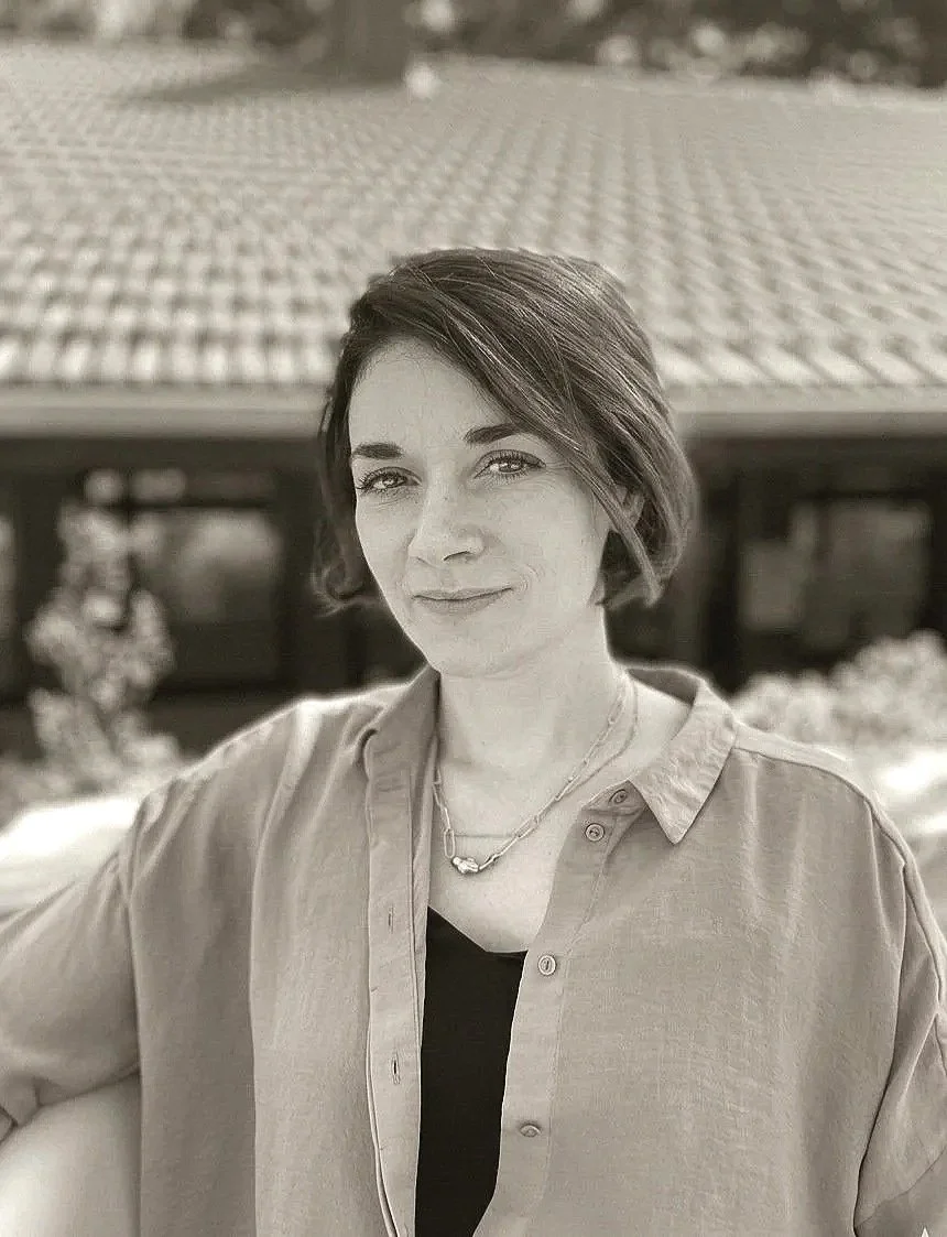 Black and white portrait of a woman with short hair, wearing a collared shirt and a necklace, standing outdoors with trees and a structural framework in the background.
