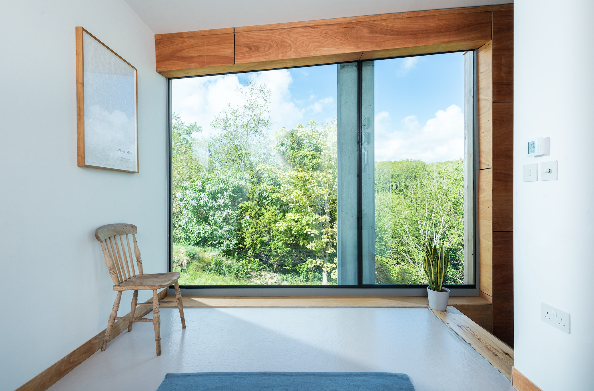 Interior view of a room with a large window showing green trees and a blue sky outside, a wooden chair, a potted plant, a framed artwork on the wall, and some electrical switches.