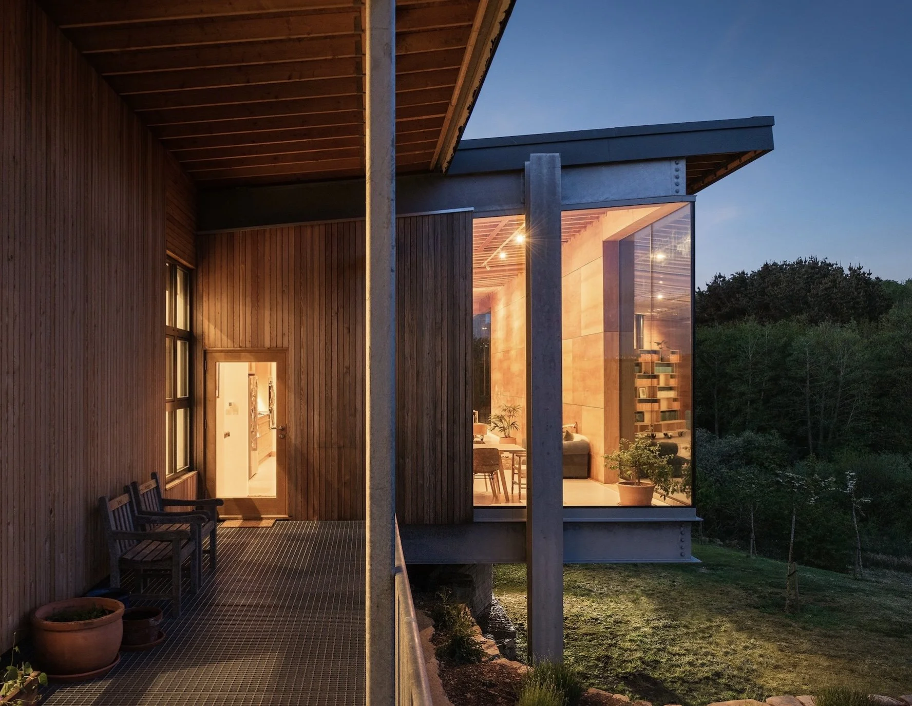 Modern house with wooden exterior and large glass windows illuminated from inside, surrounded by greenery at dusk.