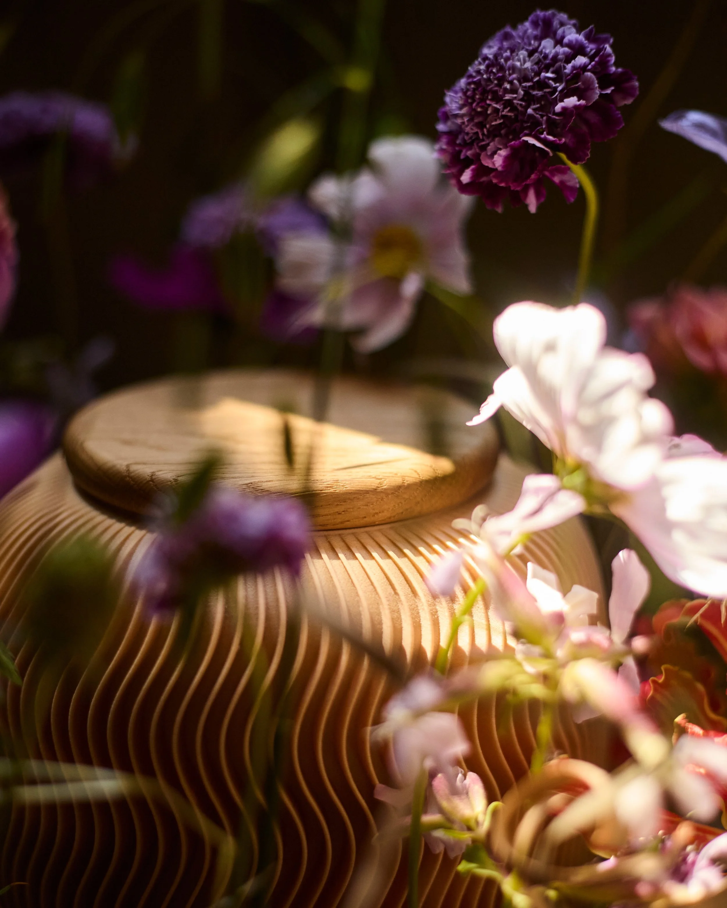 Blumenarrangement mit verschiedenen pink und lila Blumen, rund um eine Farvel Urne Nami, im Hintergrund mit dunklem Hintergrundlicht.