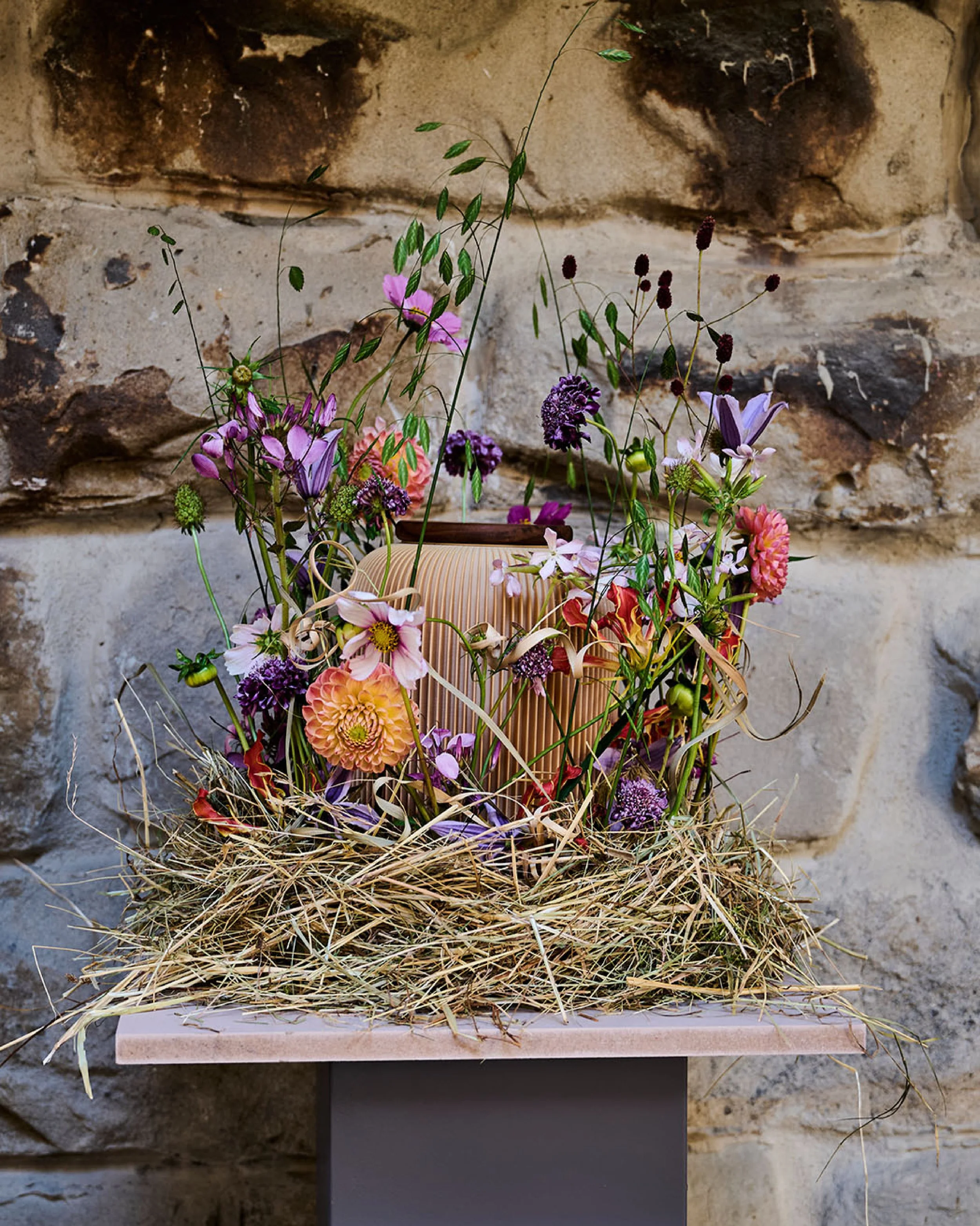 Blumenarrangement um eine Farvel Urne auf einem Tisch vor einer Steinmauer, Kryptoméer, Sonnenhut, Dahlien, Wildblumen, Trockengräser, Rinde