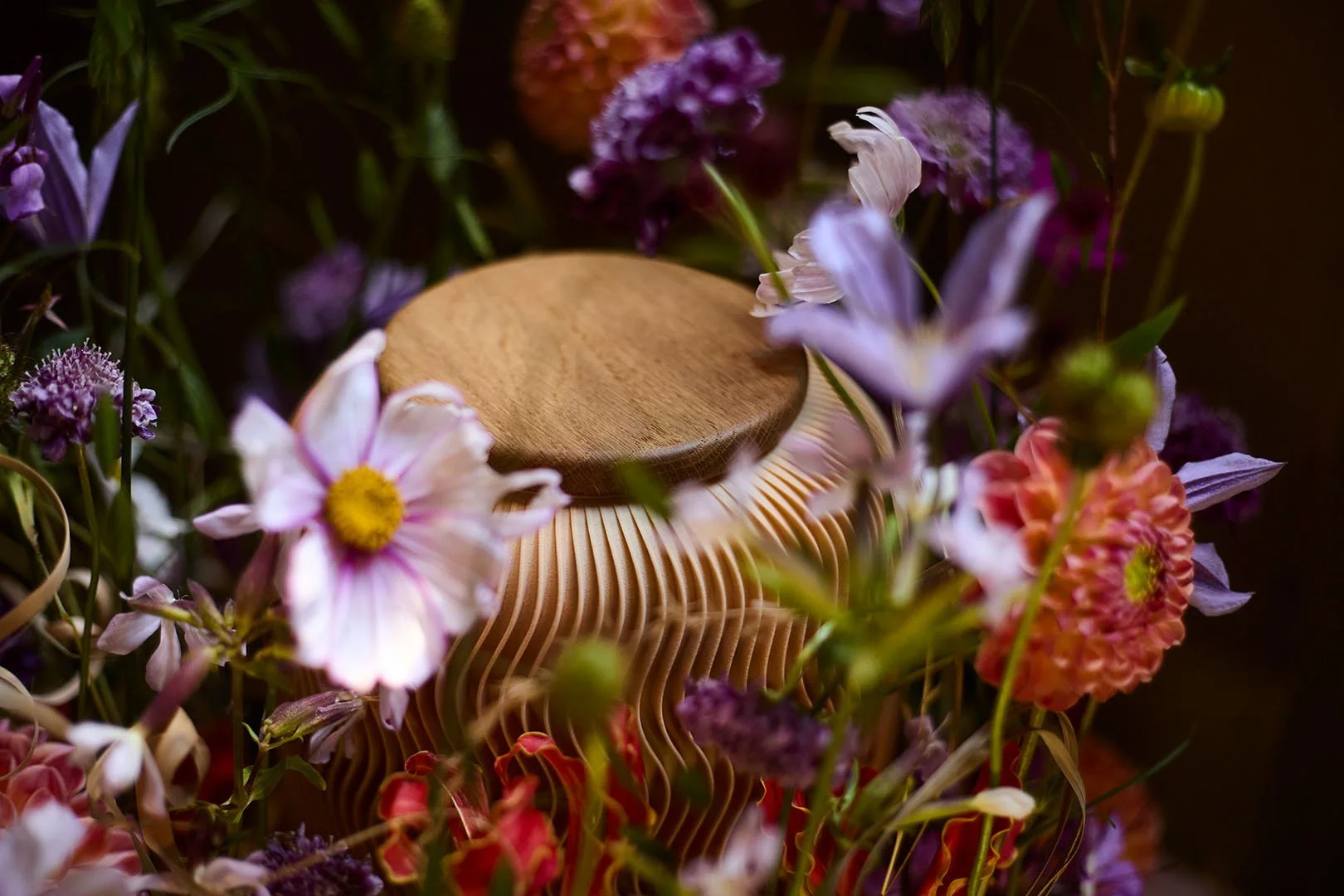 Ein runder Holzsitz auf einer Keramikvase, umgeben von bunten Blumen in verschiedenen Farben und Formen.