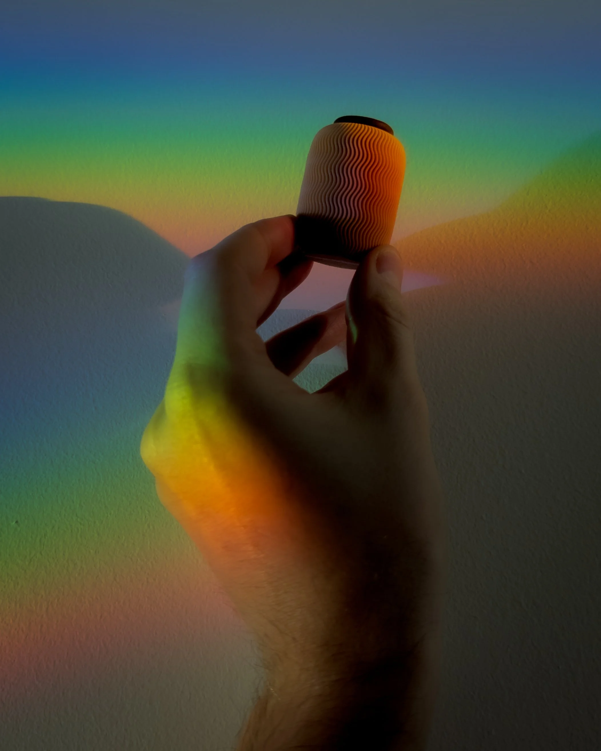 Jemand hält eine farbige, schimmernde Mikro Nami Urne von Farvel vor einer Wand, auf die Regenbogenfarben projiziert werden.