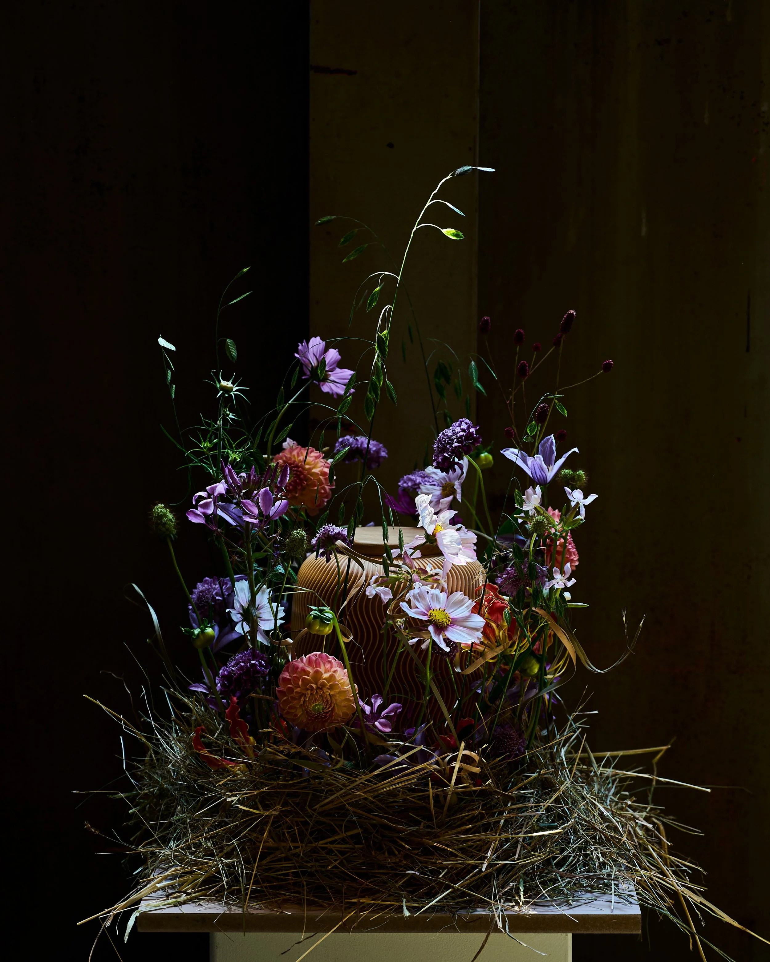 Urne Minka auf einer erhöhten Plattform, umgeben von Stroh, mit unterschiedlichen bunten Blumen vor dunklem Hintergrund.