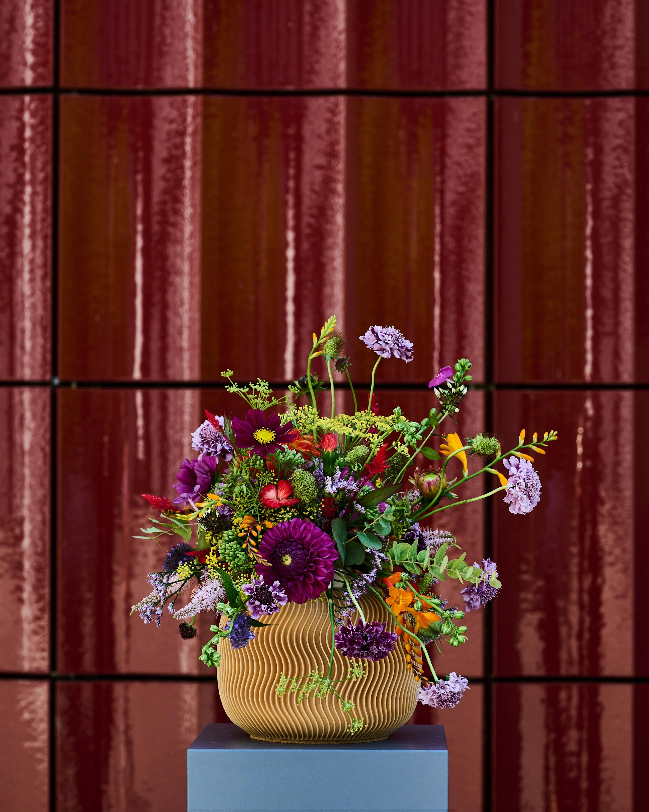 Blumenstrauss mit bunten Blumen in einer modernen Mini Urne Nami auf einem blauen Podest, vor einem roten Ziegelsteinhintergrund.