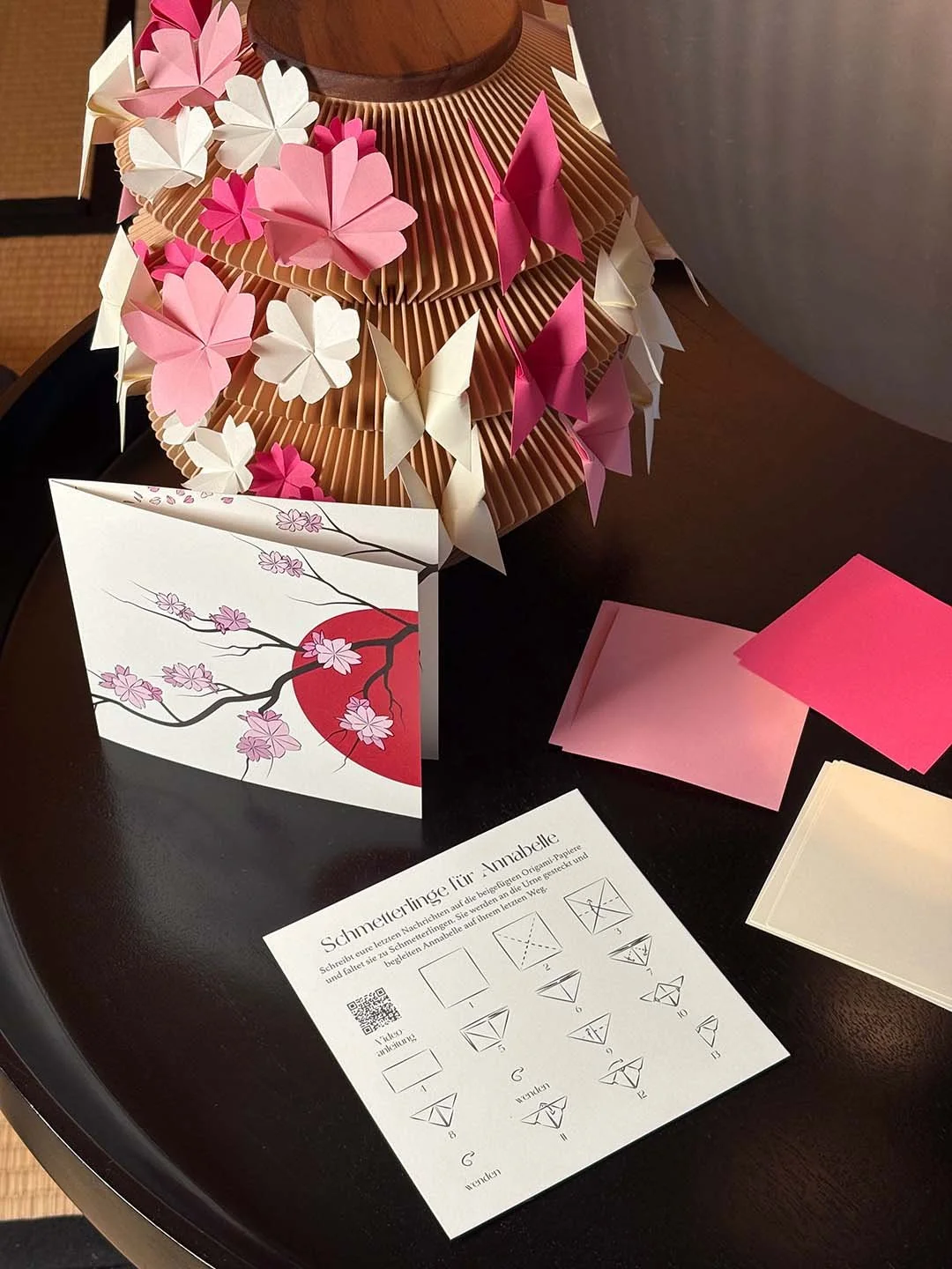 Origami-Schmuck aus rosa, weißen und pinken Papierblumen und Schmetterlingen, dazu eine Bastelanleitung für Origami-Anhänger und farbige Papptücher auf einem Tisch.