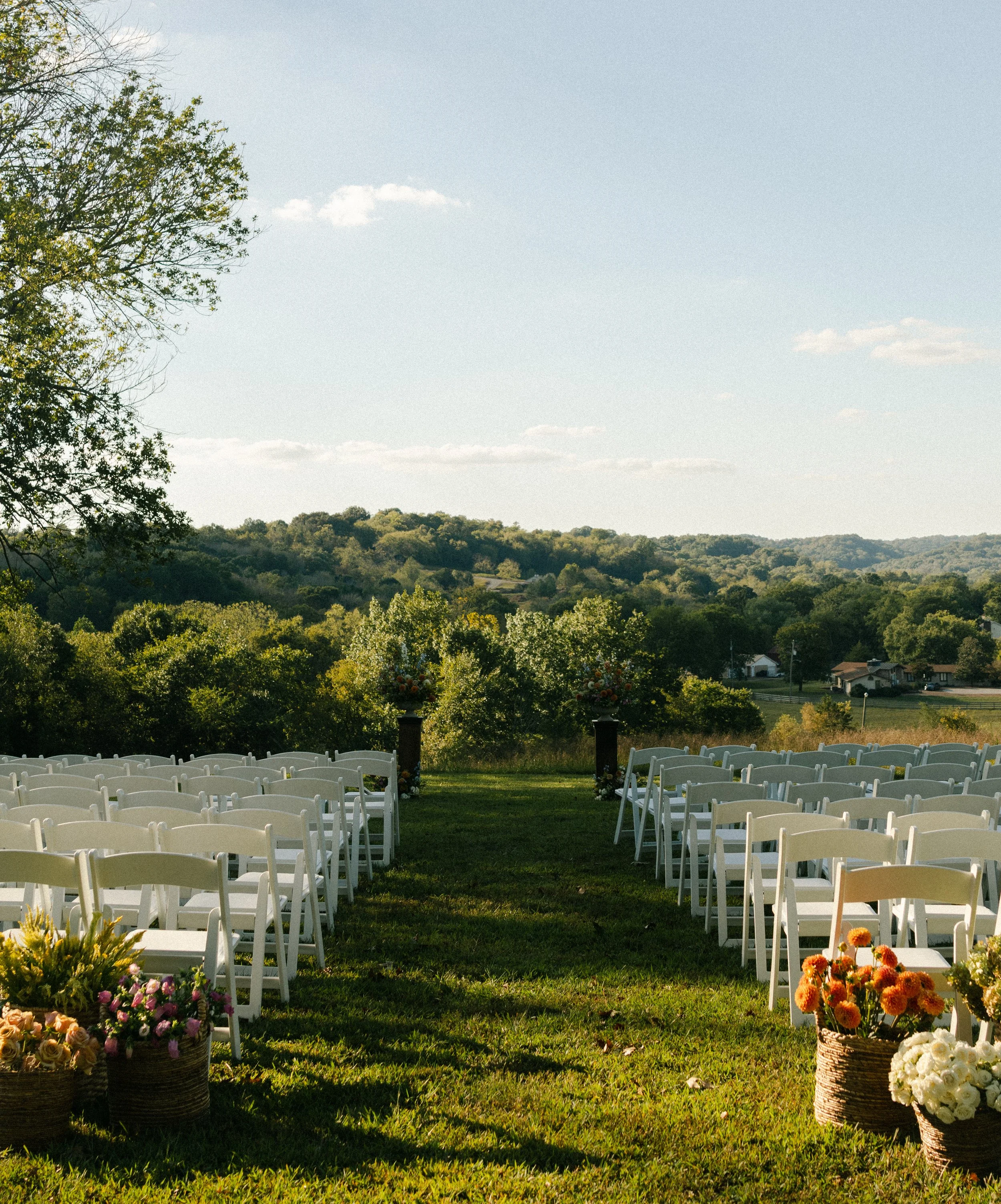 Outdoor wedding ceremony setup with white chairs in rows, floral arrangements in baskets, and a natural landscape with trees and rolling hills under a partly cloudy sky.