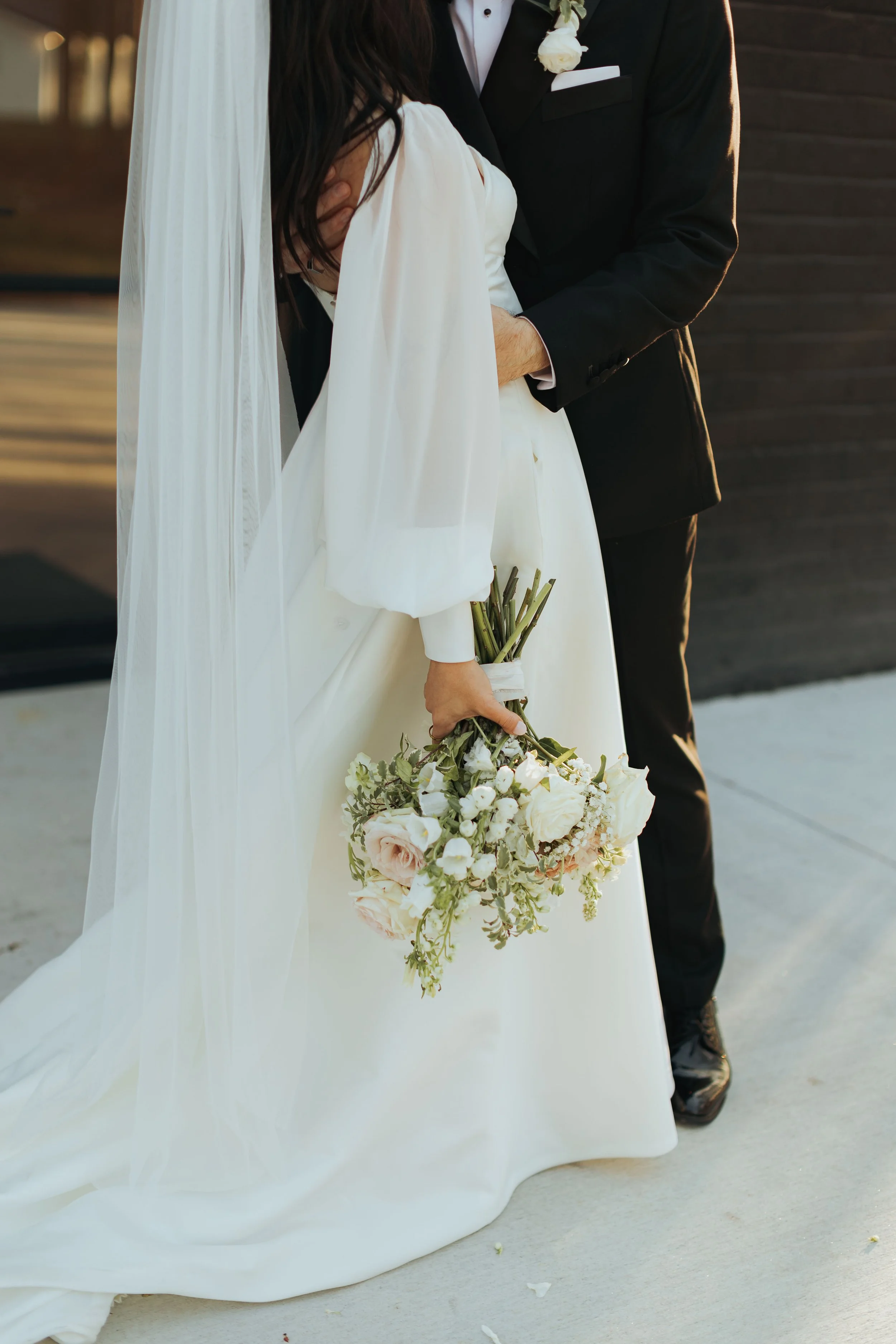 Close-up of a bride holding a bouquet of white and pale pink flowers, wearing a white wedding gown and veil, standing next to a groom in a black tuxedo with a white boutonniere.
