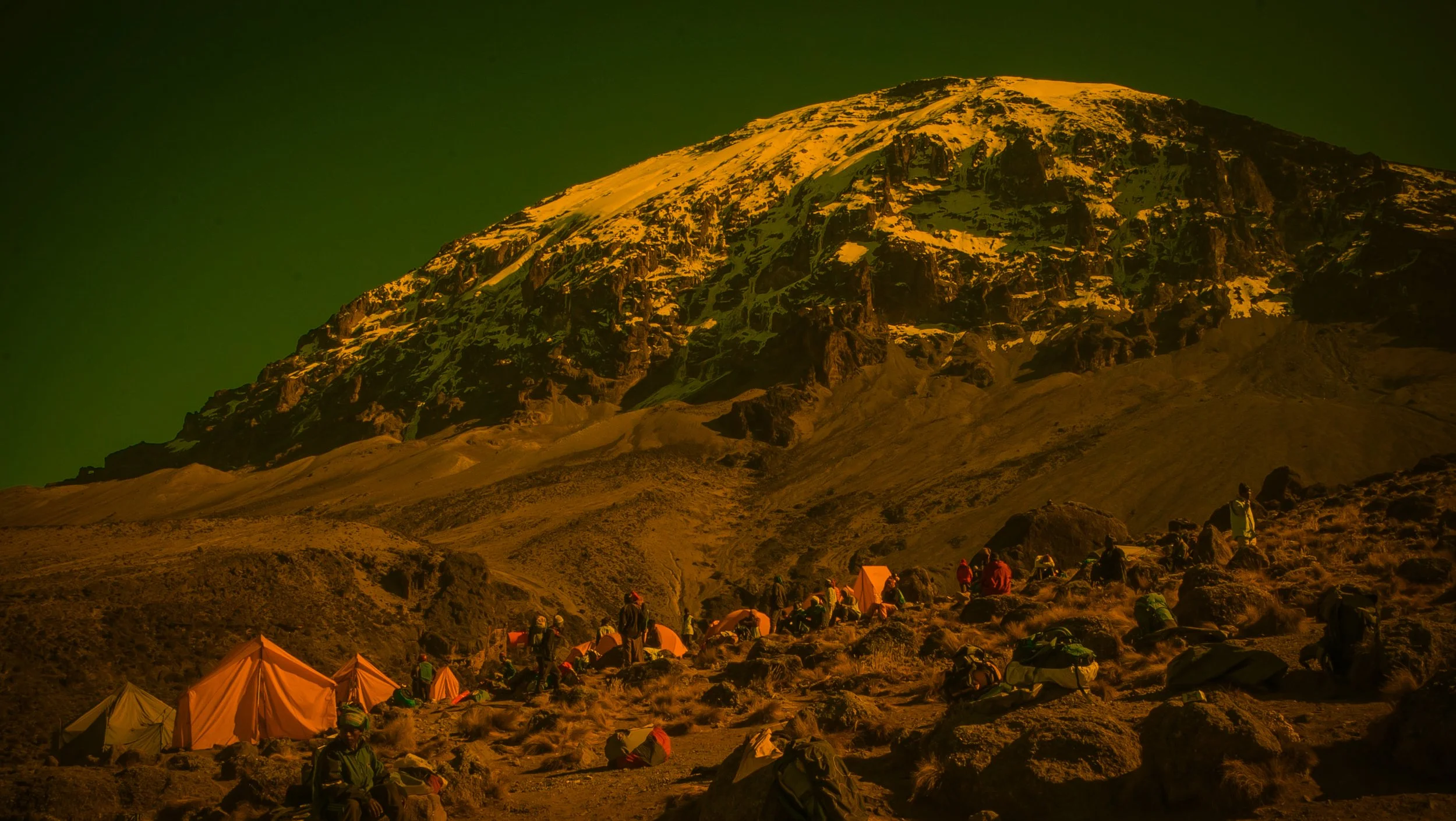 Camp setup on a rugged mountain landscape with a snow-capped volcano in the background during sunset or sunrise.