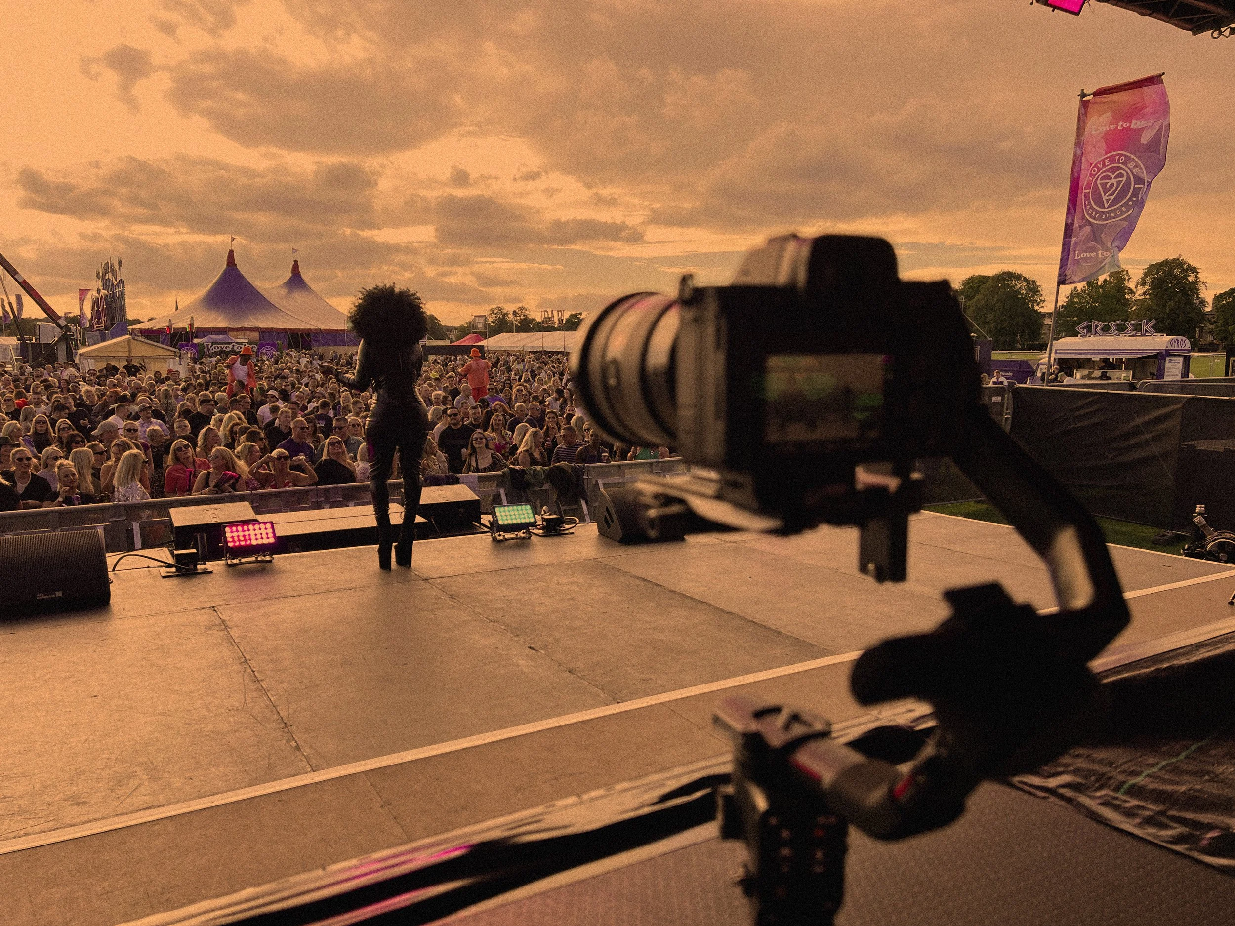 On stage at a concert, camera in foreground, performer and crowd in background, with colorful festival tents and flags, during sunset.