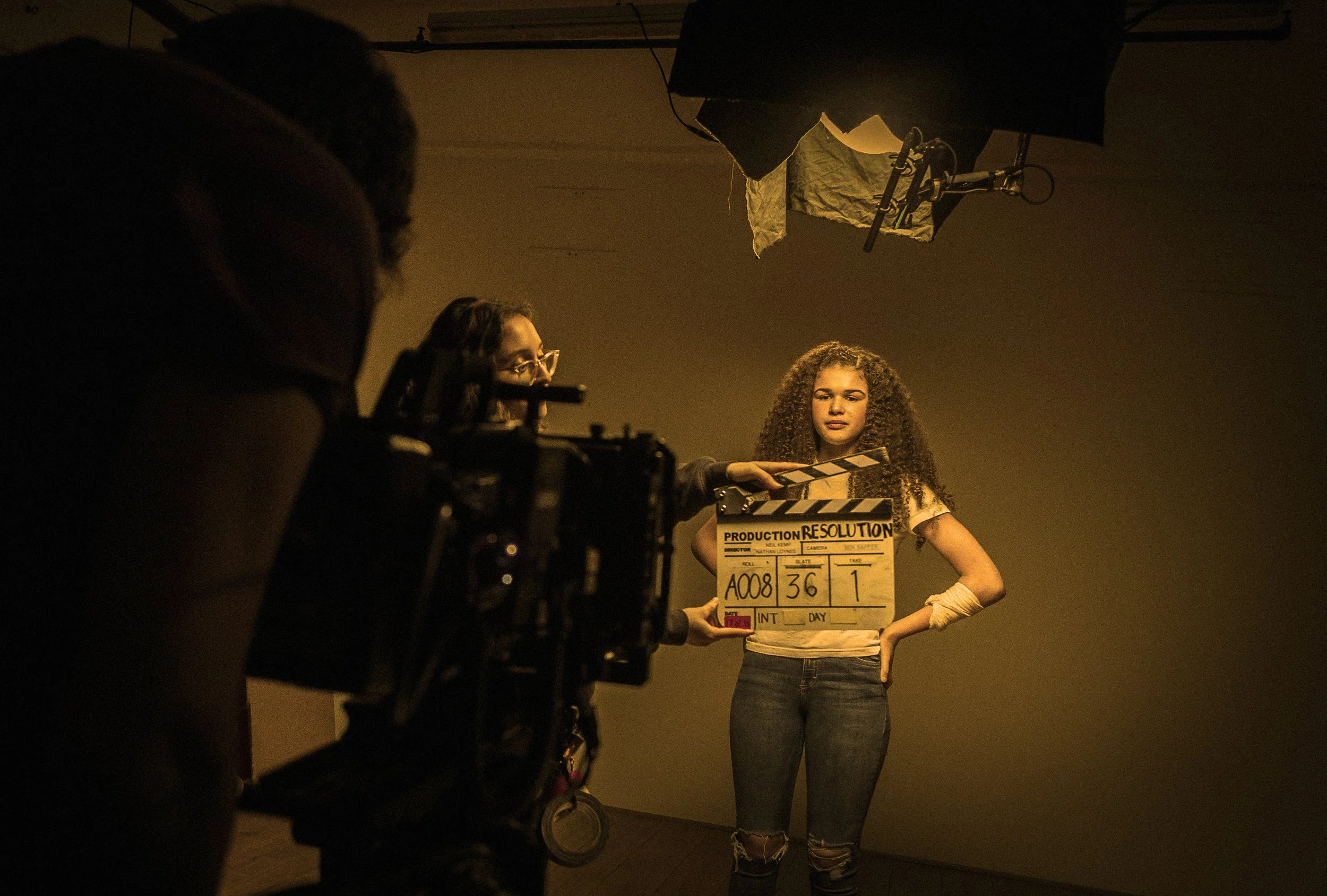 A young woman with curly hair is standing in front of a camera, holding a clapperboard on a film set. Two crew members are assisting her, one of whom is visible on the left side of the image. The scene is lit with warm studio lighting.