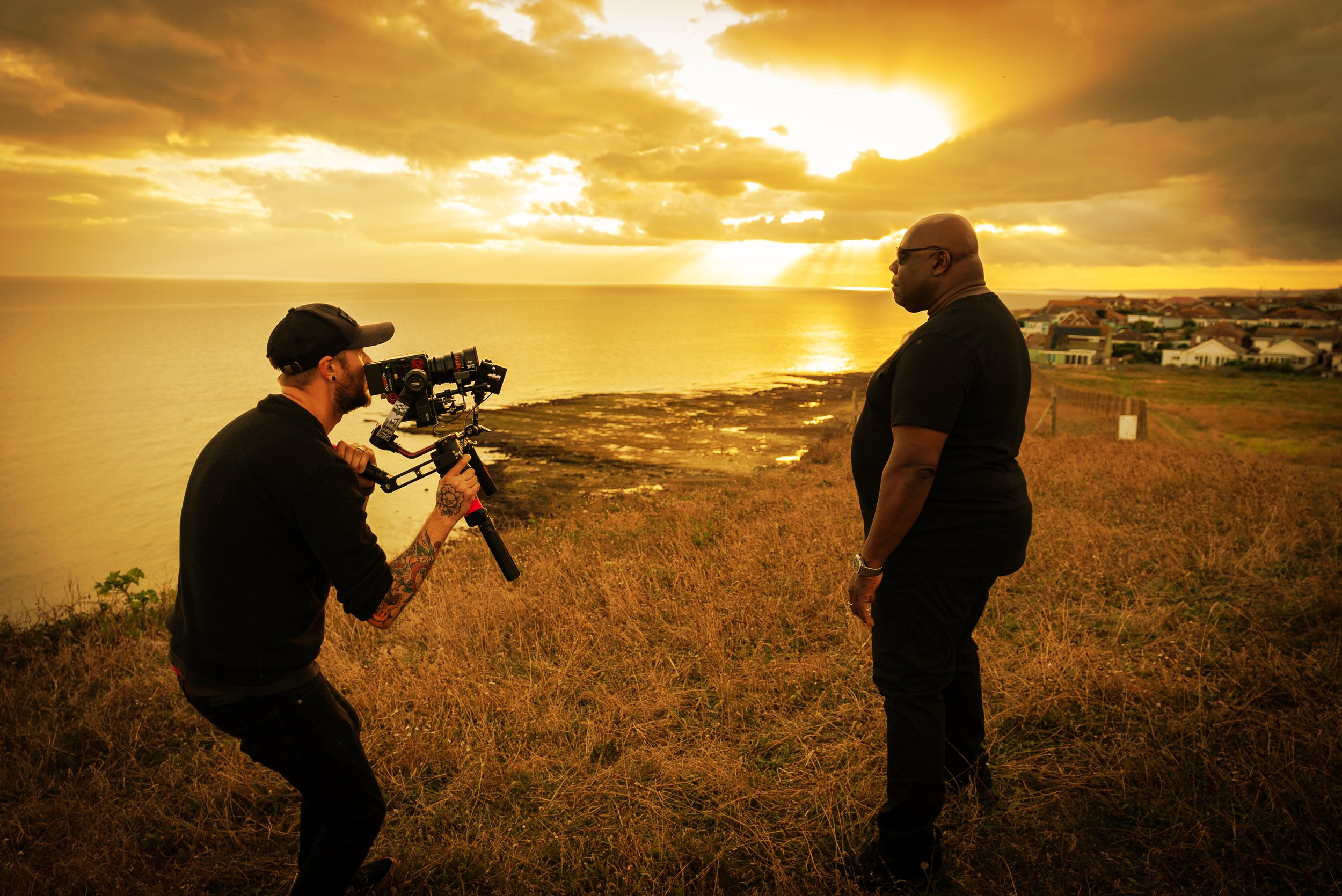 A man filming another man on a grassy shoreline during sunset, with houses and a body of water in the background.