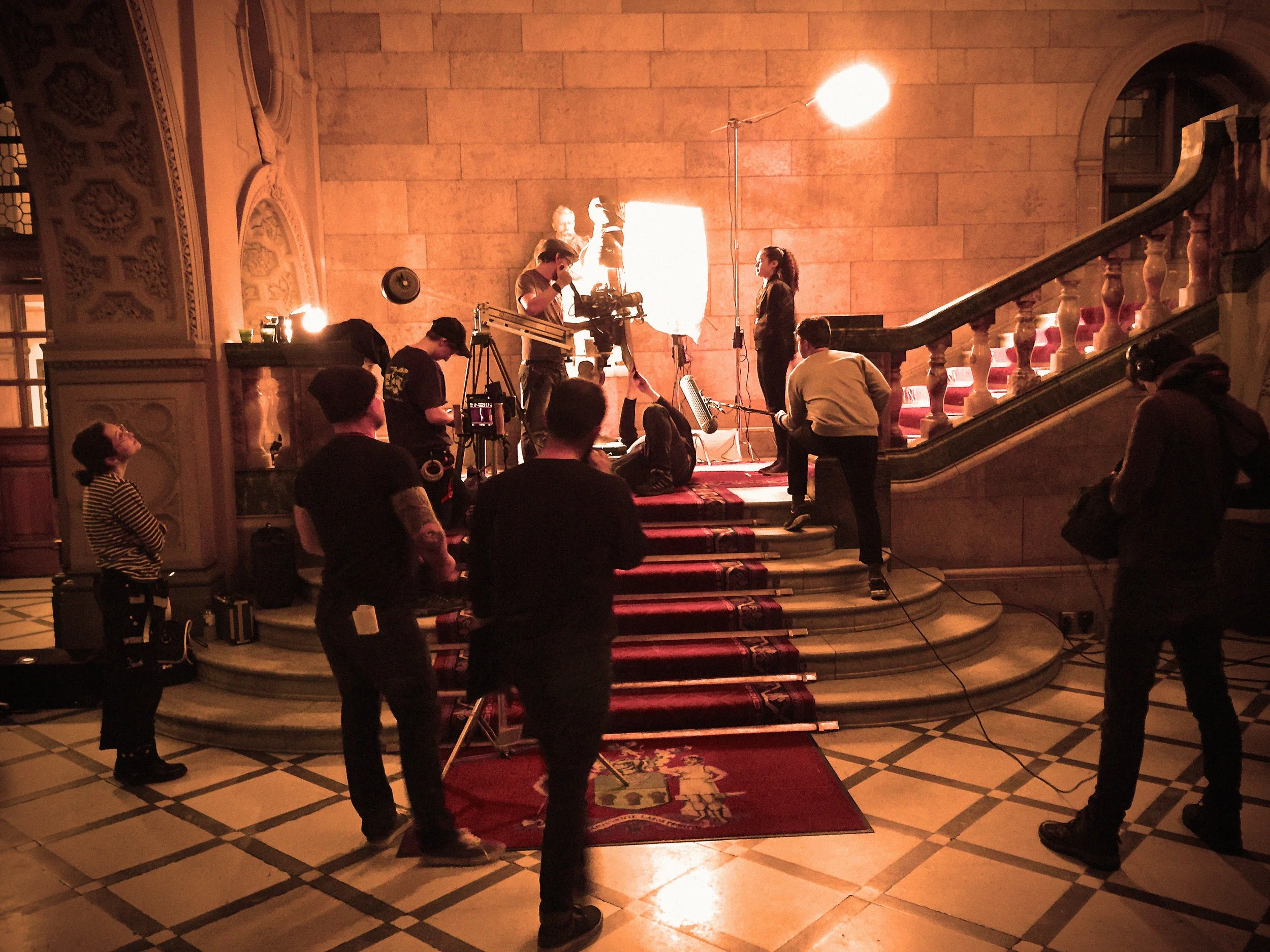 Film crew setting up and filming a scene on a staircase inside a grand building with ornate decor, warm lighting, and a red carpet in the foreground.