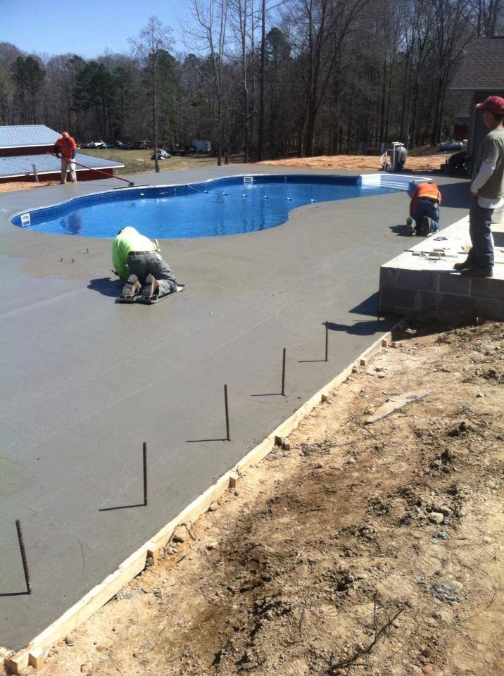 Construction workers installing a concrete patio around an in-ground swimming pool in a wooded area.