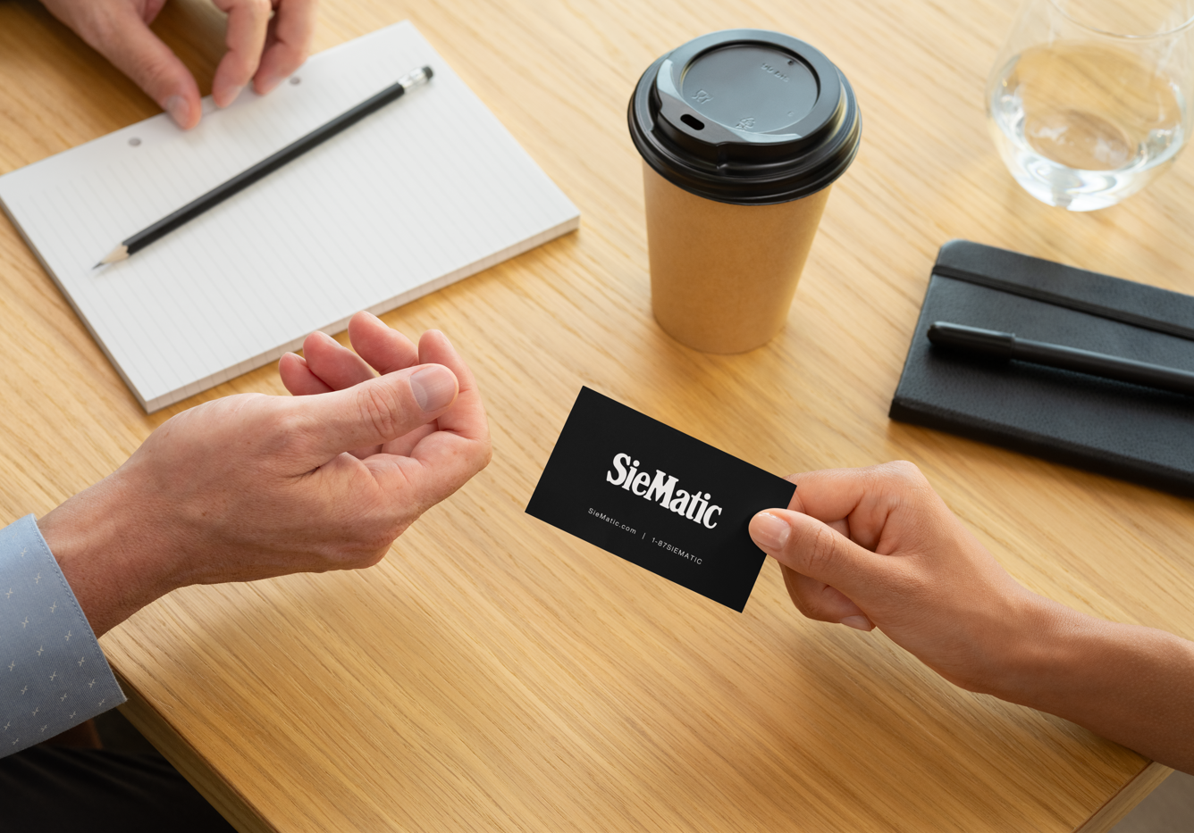 A person exchanging a black business card with the text 'SieMatic' on it, during a meeting or discussion at a wooden table.