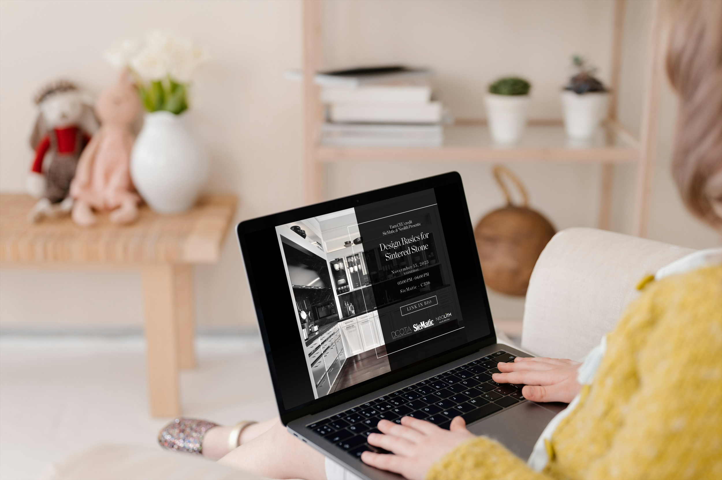 Person sitting on a beige chair using a laptop, showing a design presentation titled 'Design Basics for Sintered Stone' on the screen. In the background, there is a wooden table with stuffed animals and a white vase with flowers, and a shelving unit with books and decorative items.