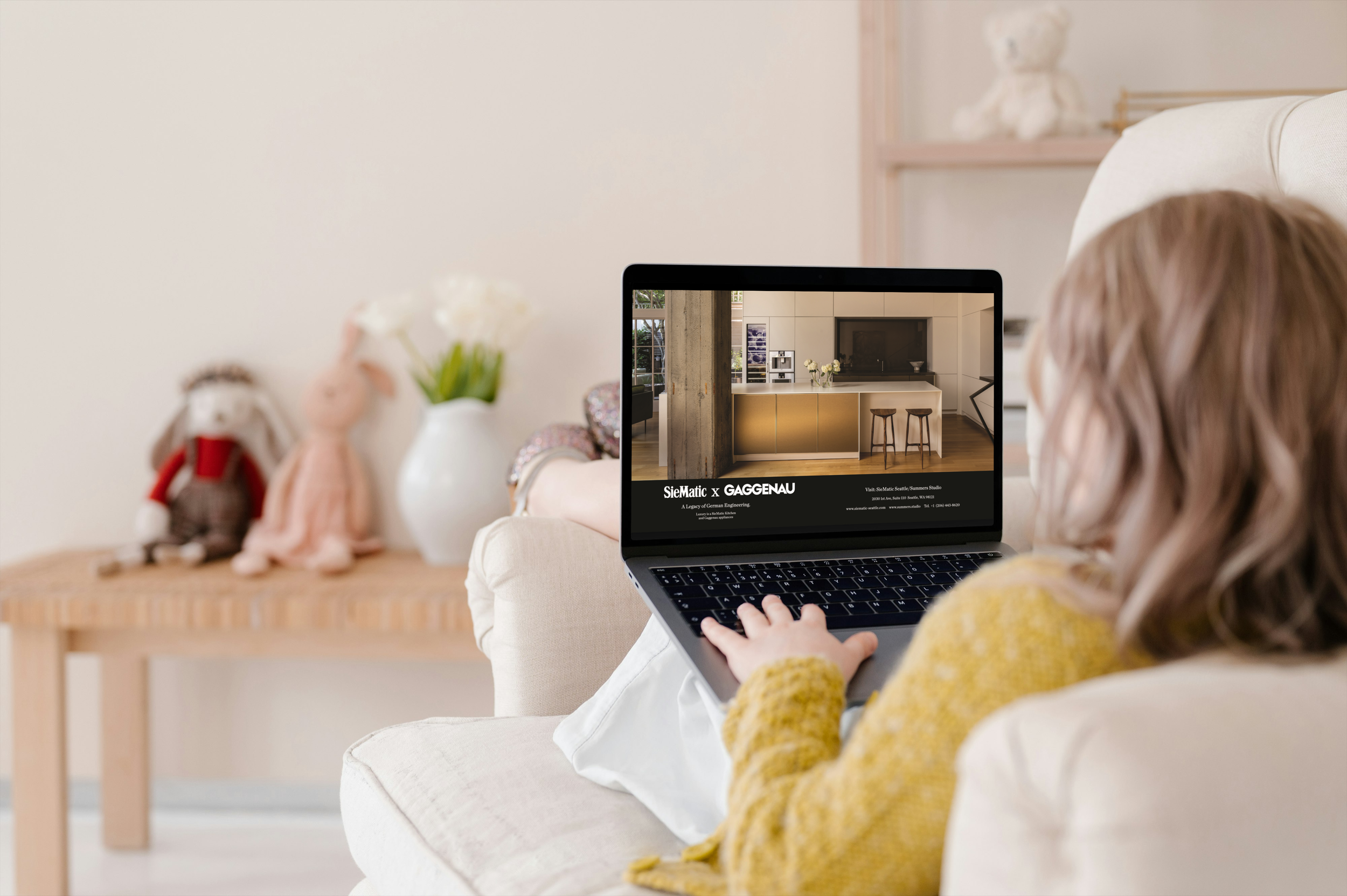 A woman sitting on a beige armchair uses a laptop showing interior design plans for a kitchen renovation.