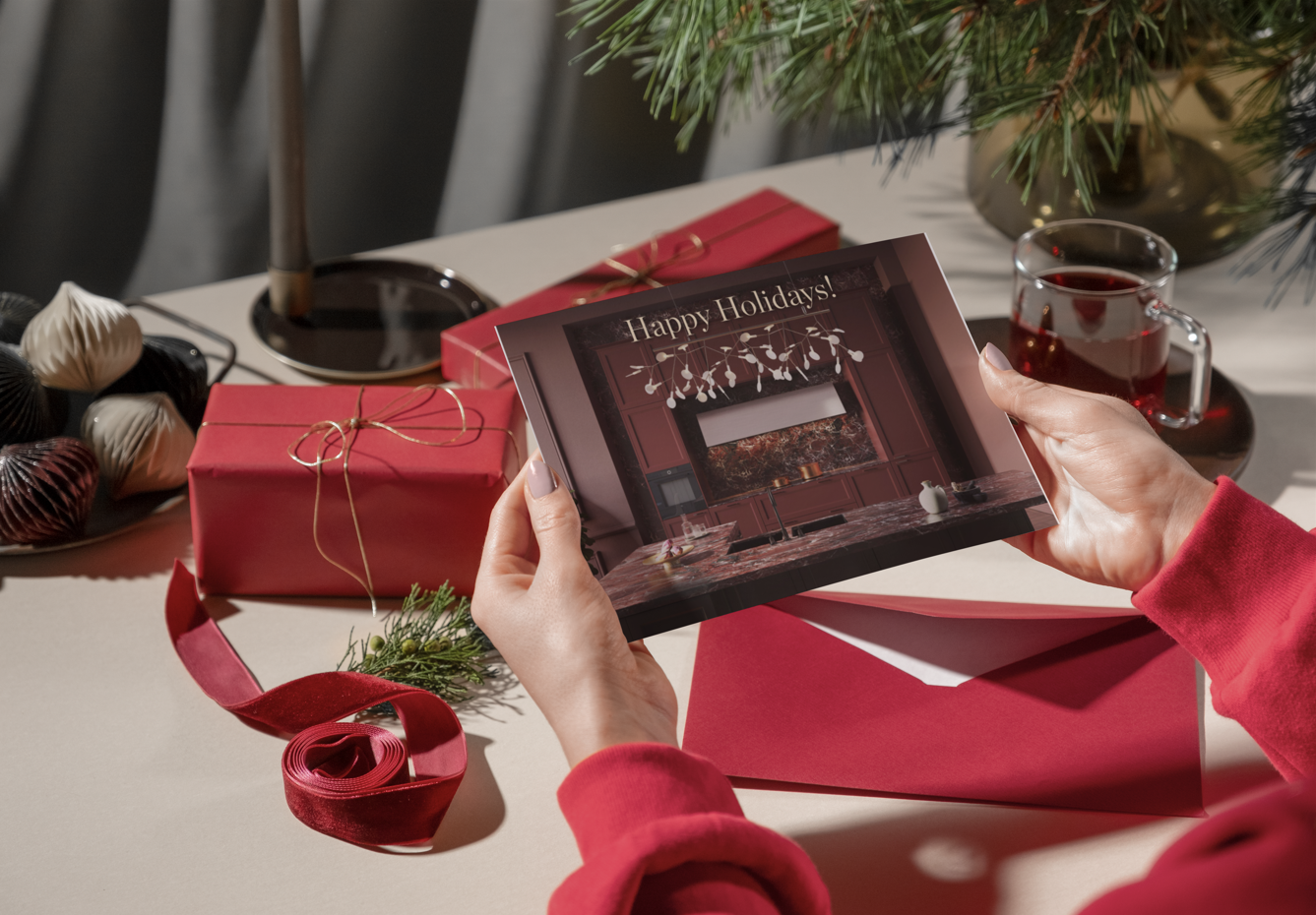 Person holding a holiday greeting card that says "Happy Holidays!" with a kitchen scene on it, surrounded by wrapped presents, Christmas ornaments, a red ribbon, and a glass cup of a red beverage on a table decorated for Christmas.